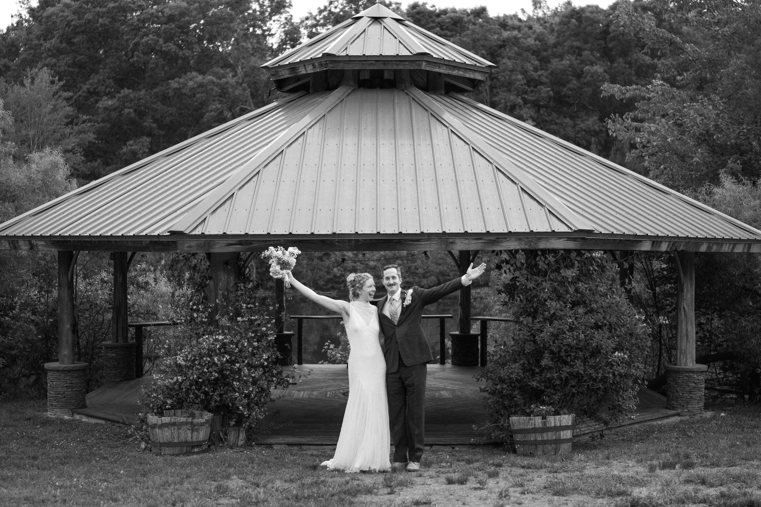 A bride and groom are standing under a wooden gazebo with trees in the background. The bride is wearing a white wedding dress and holding a bouquet, while the groom is dressed in a dark suit. They are smiling and celebrating.