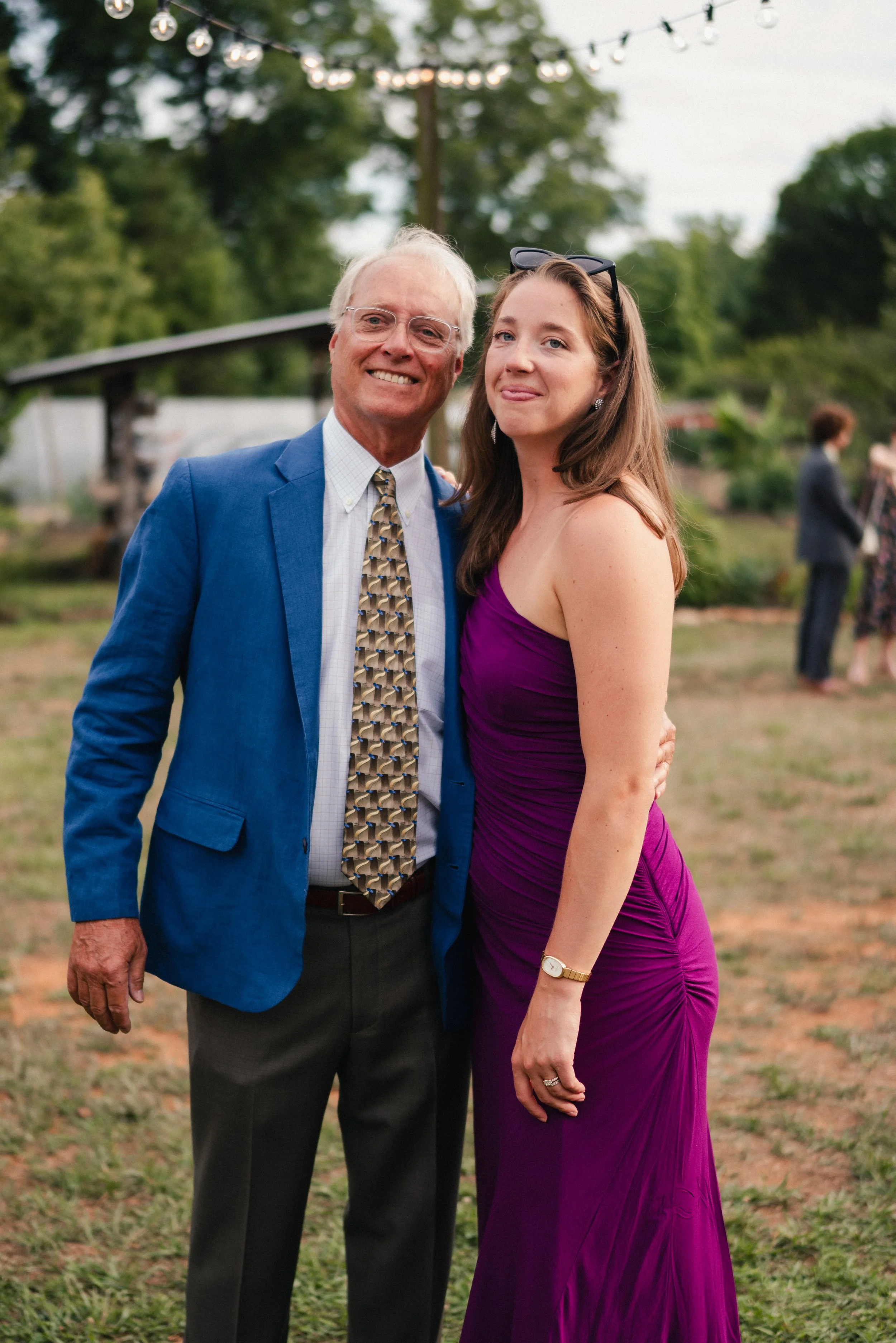 An older man in a blue blazer, white shirt, and patterned tie standing next to a woman in a purple dress with sunglasses on her head, both smiling at an outdoor event with string lights overhead and other people in the background.