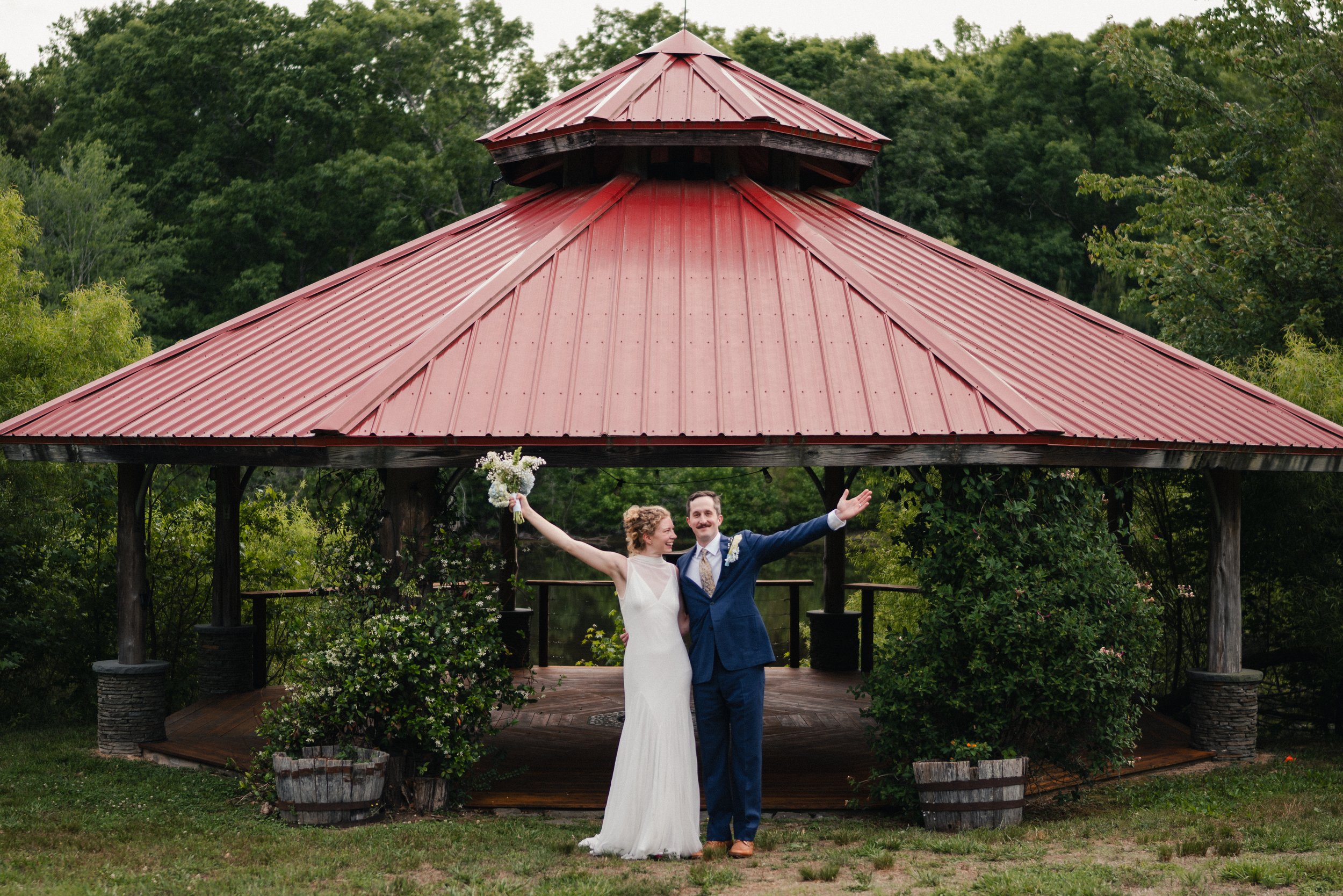 A bride and groom celebrating outdoors under a red metal pavilion, near greenery and trees, with their arms raised.