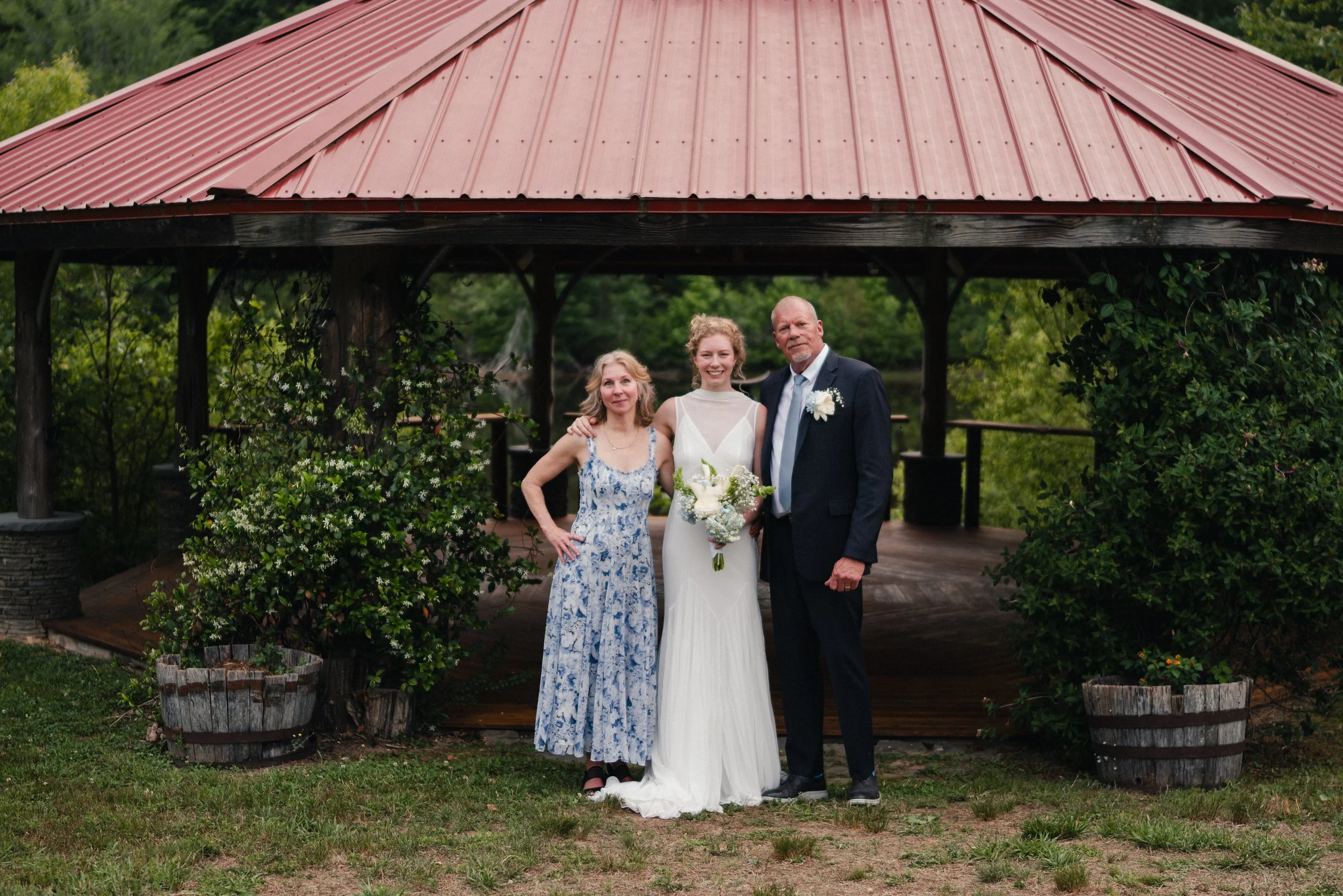 A bride in a white wedding dress with a bouquet, standing between her parents, in front of a red metal gazebo, on a grassy area with greenery.