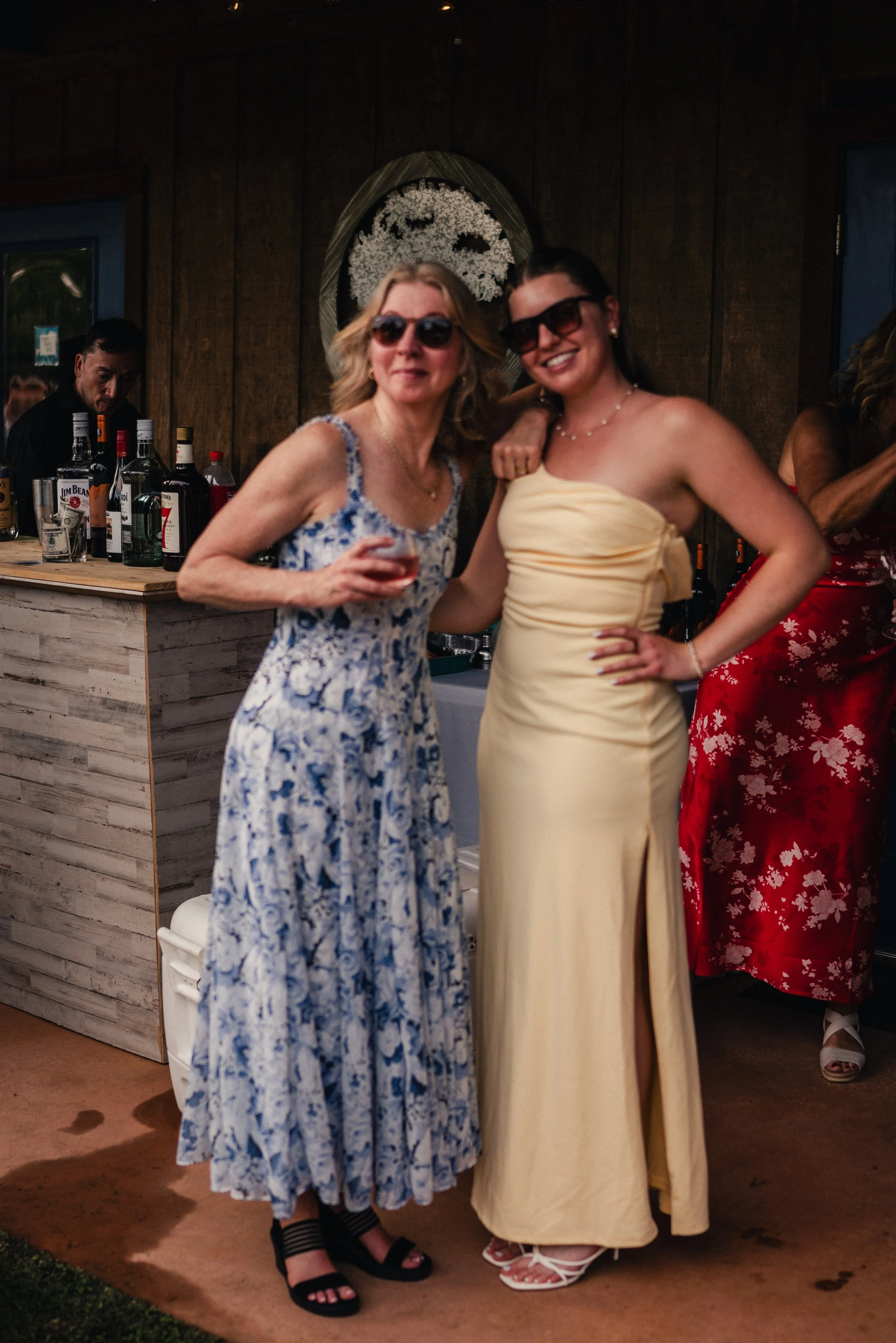 Two women wearing summer dresses and sunglasses pose together at an indoor event, with a bar and bartender in the background.