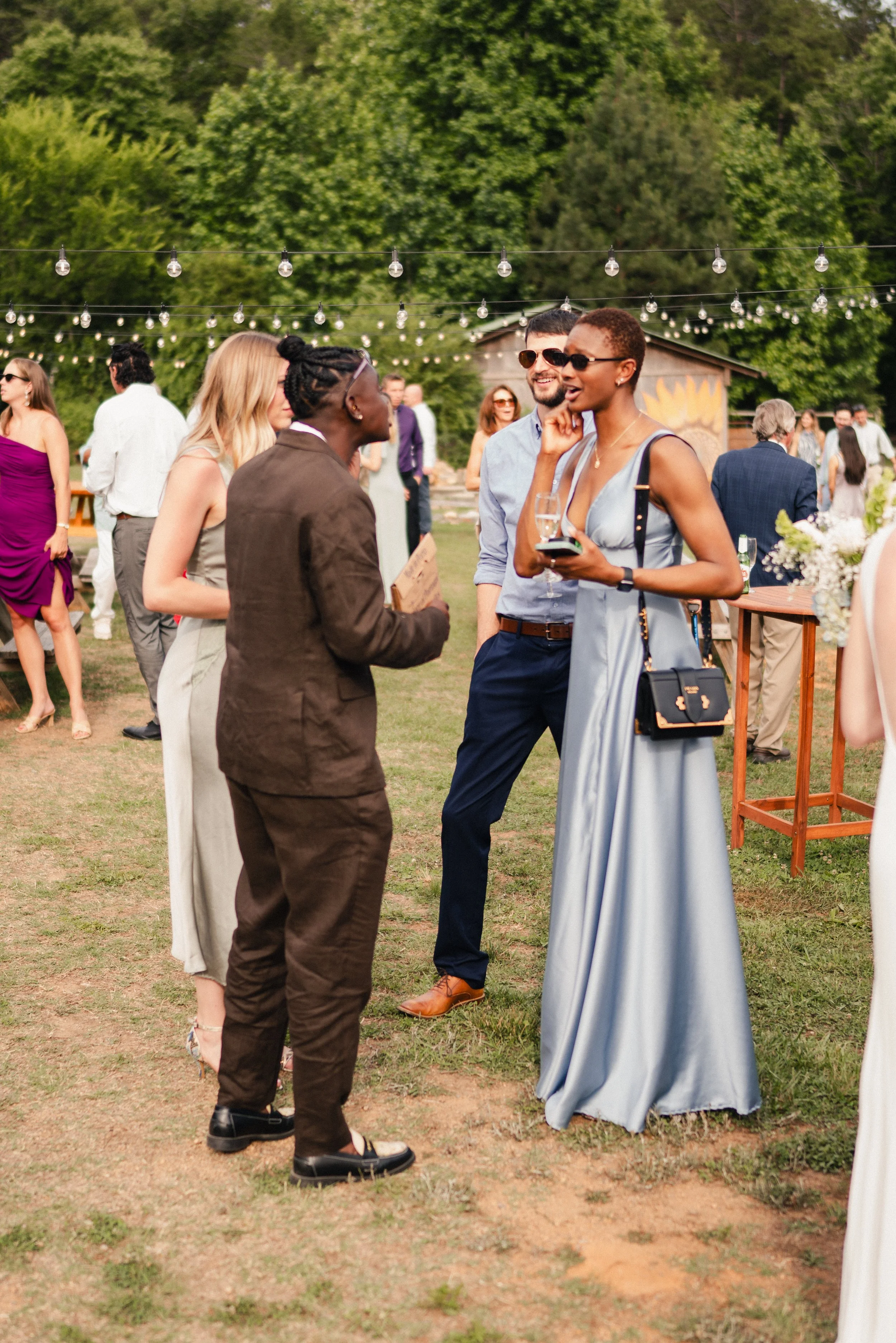 People in formal attire at an outdoor event under string lights, with lush green trees in the background.