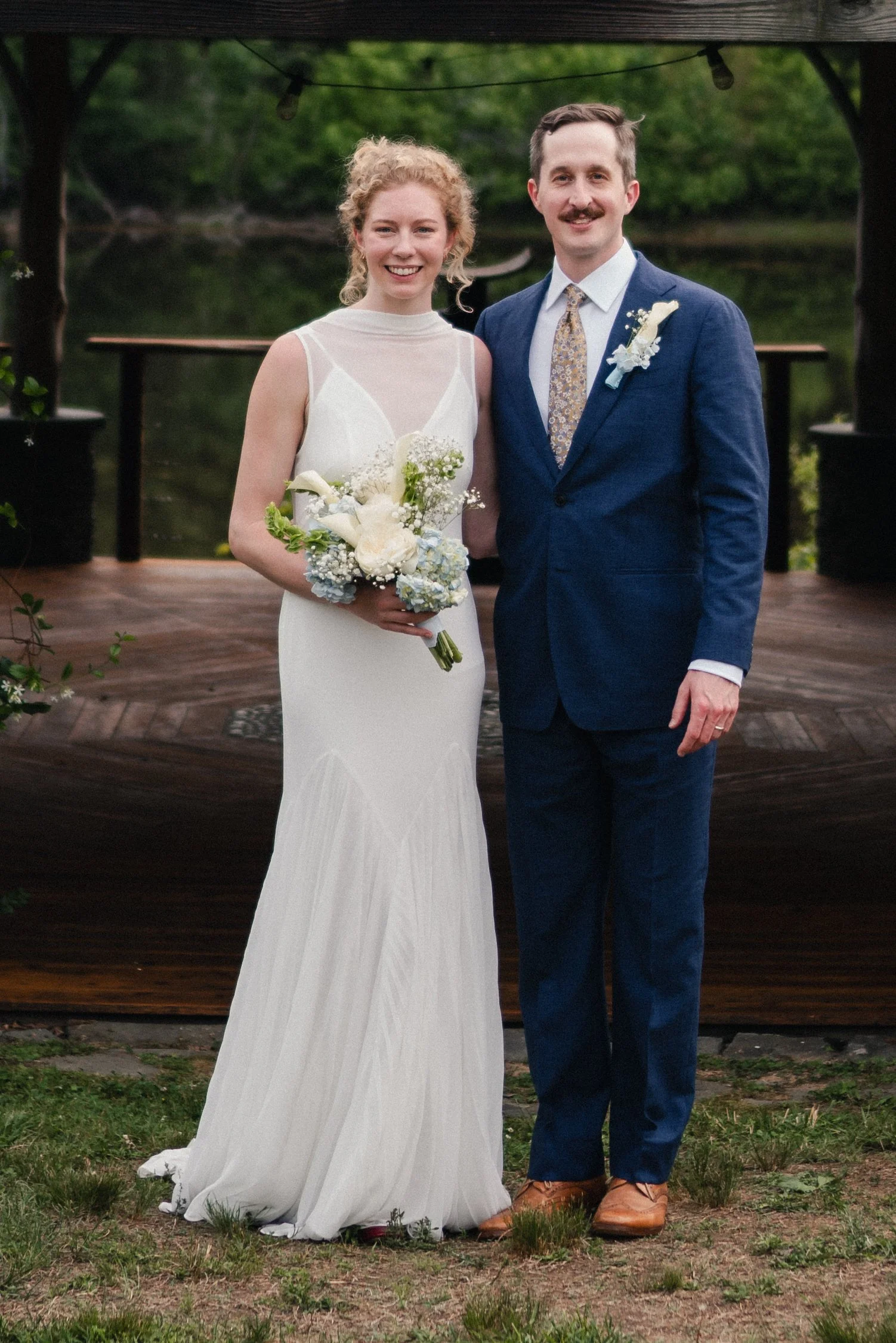 A bride and groom stand together outdoors on a wooden deck, smiling for a photo. The bride wears a white wedding gown and holds a bouquet of white and pale blue flowers. The groom wears a navy blue suit with a floral tie and a white boutonniere. The 