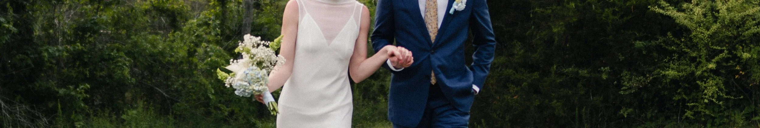 A bride and groom holding hands while walking outdoors in a wooded area.