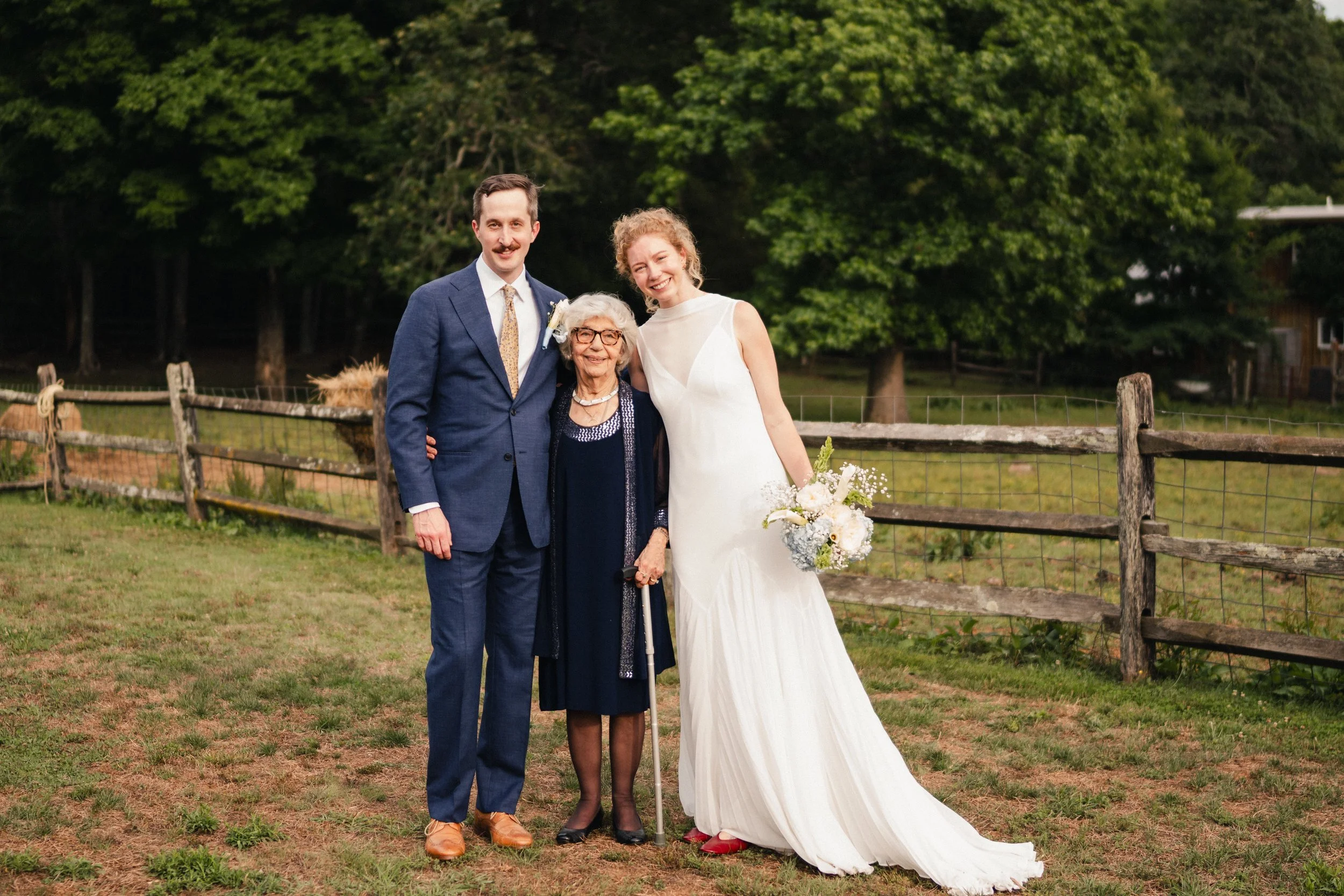 A bride in a white wedding dress standing outdoors with a man in a blue suit and an elderly woman in dark clothing, all smiling for a photo on a grassy area with a wooden fence and green trees in the background.