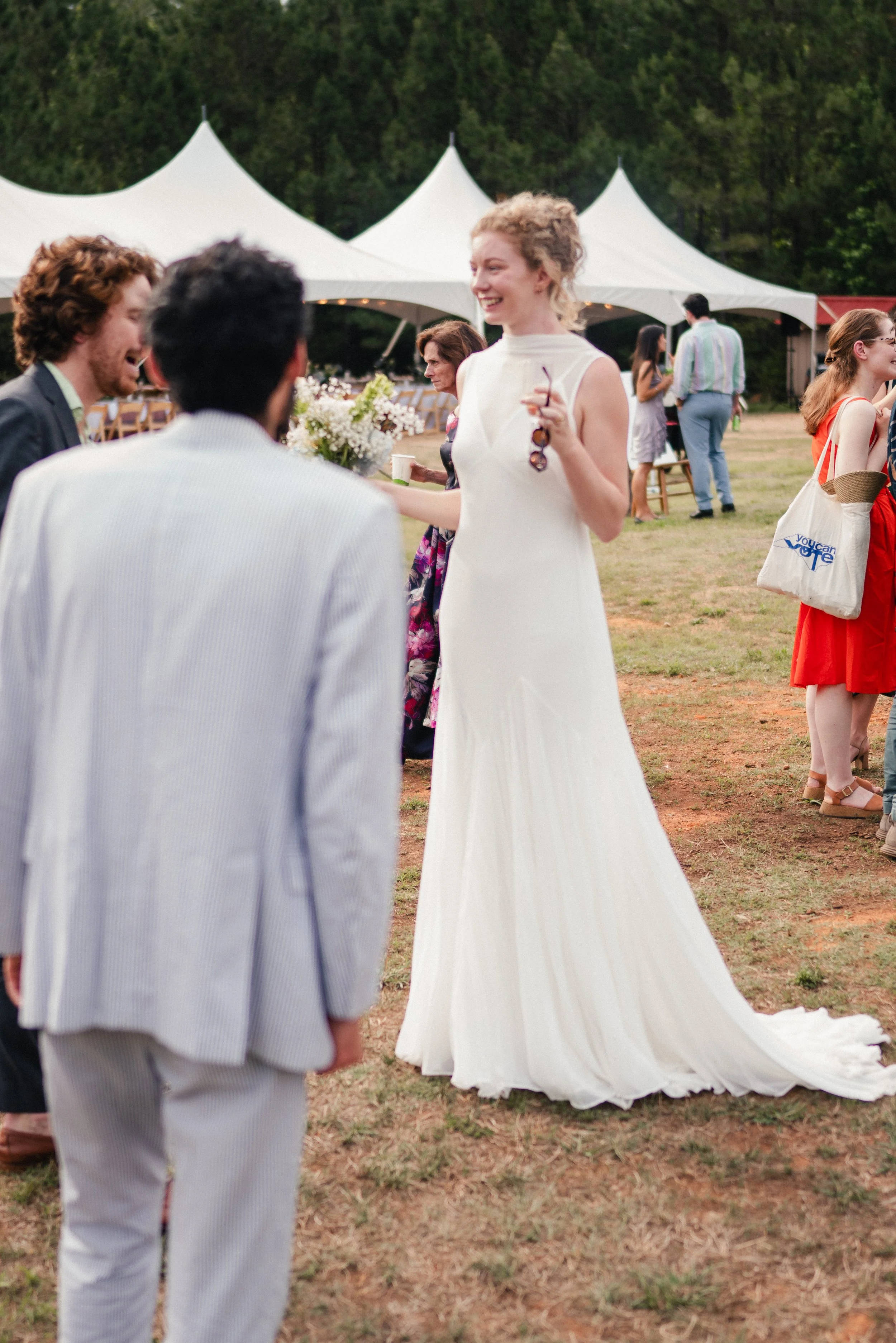 People at an outdoor event, including a woman in a white dress holding keys and glasses, surrounded by others in casual and semi-formal attire, with white tents and trees in the background.