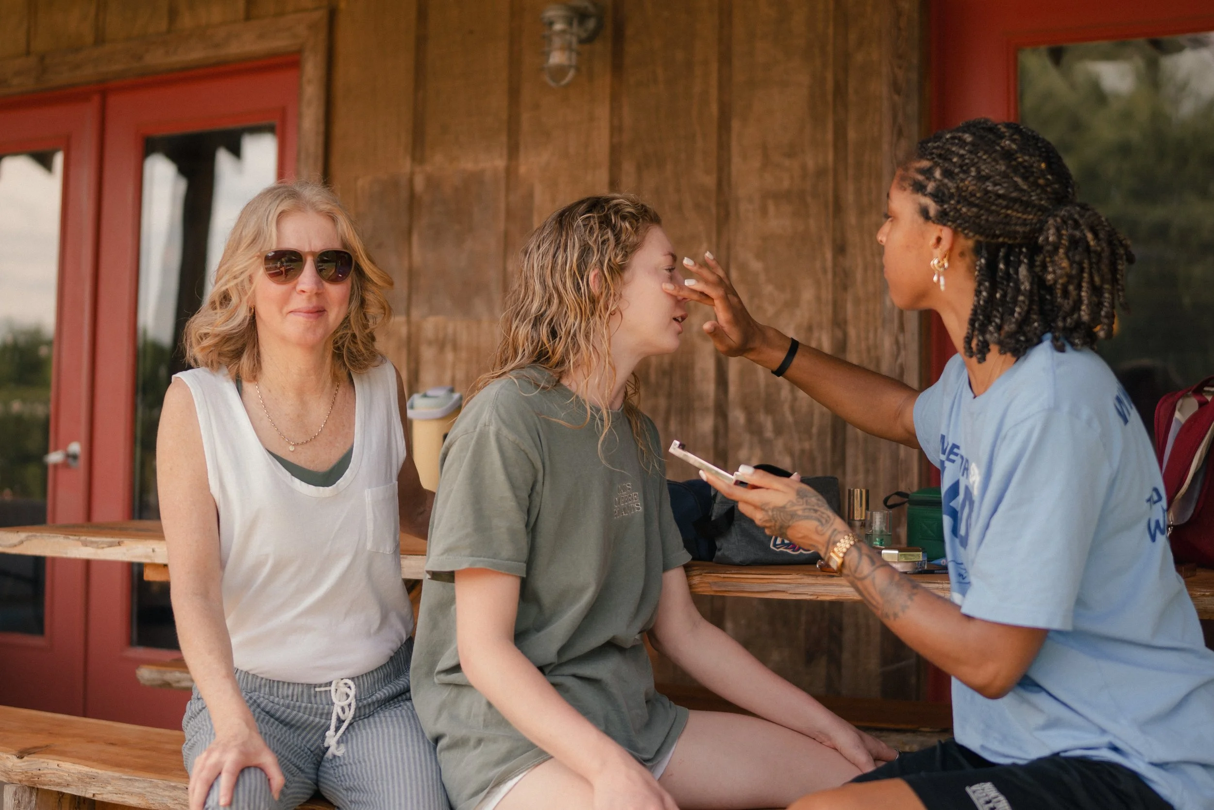 A woman with blonde hair and sunglasses standing beside a young woman with curly blonde hair, who is sitting, as a woman with braids and earrings touches her nose. The scene takes place outdoors with wooden background and windows.