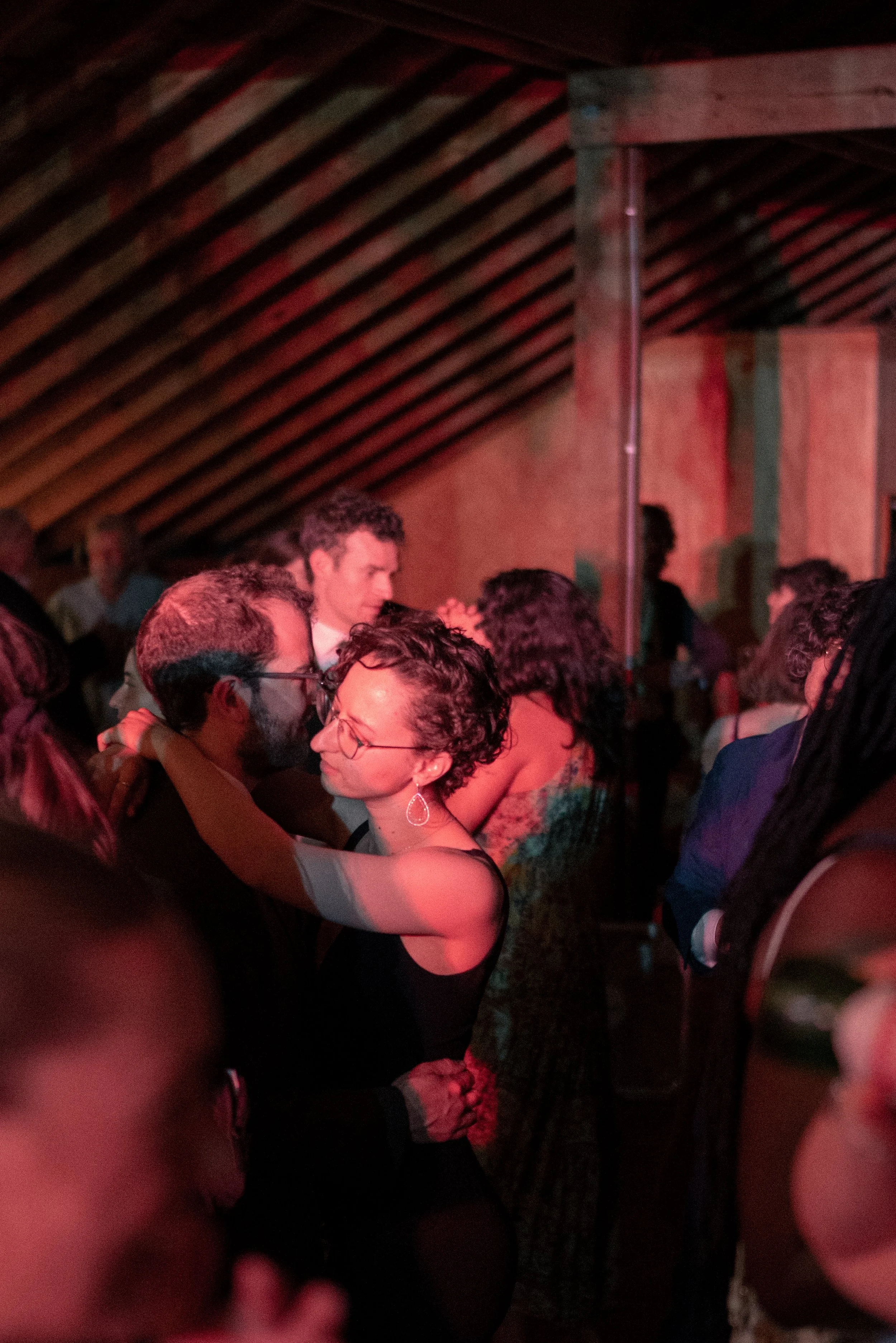 People dancing closely together at a social event in a dimly lit room with a red hue and sloped wooden ceiling.