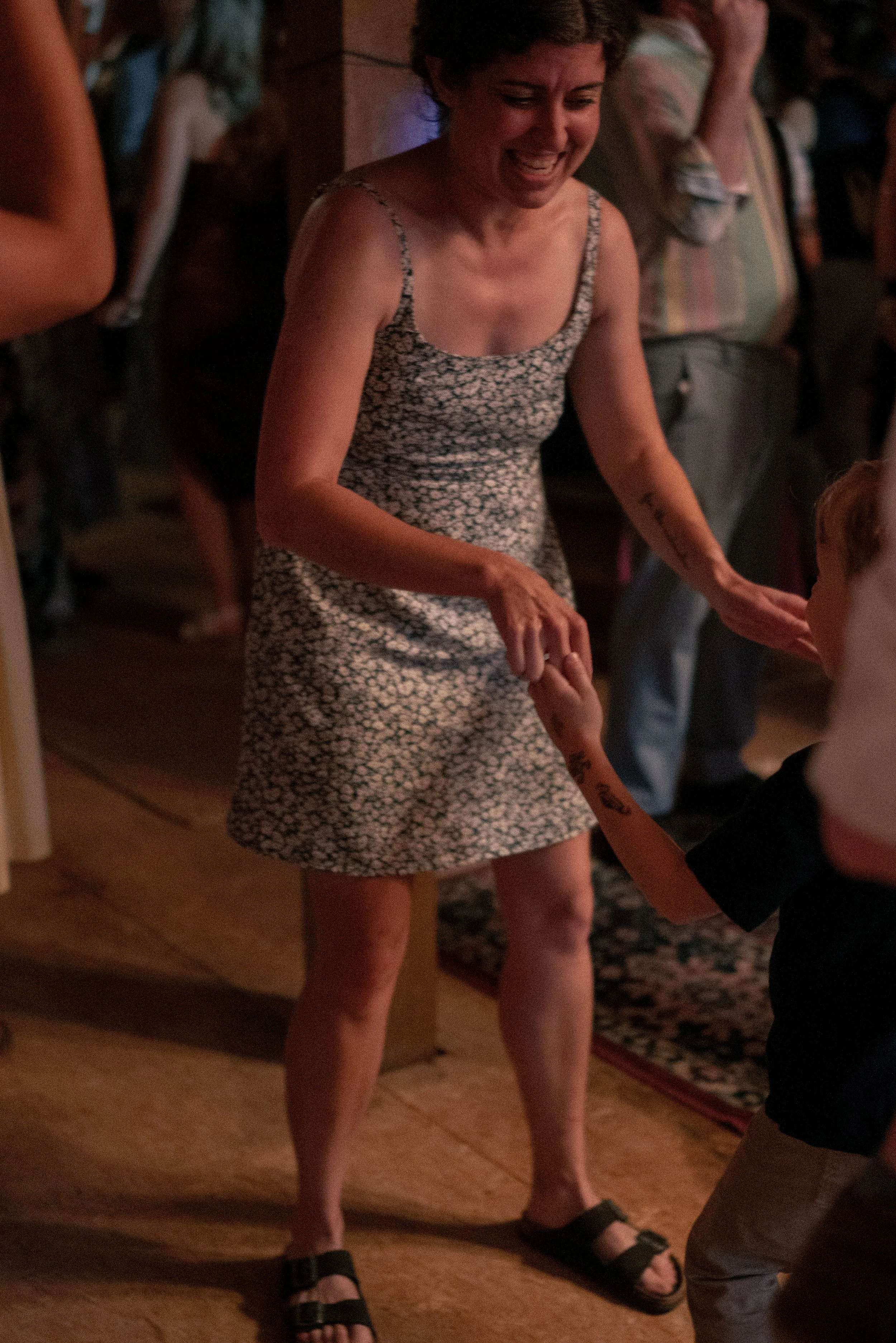 A woman with short hair wearing a sleeveless, patterned dress, smiling and holding hands with a young child in a dark outfit, in a room with a wooden floor and other people in the background.