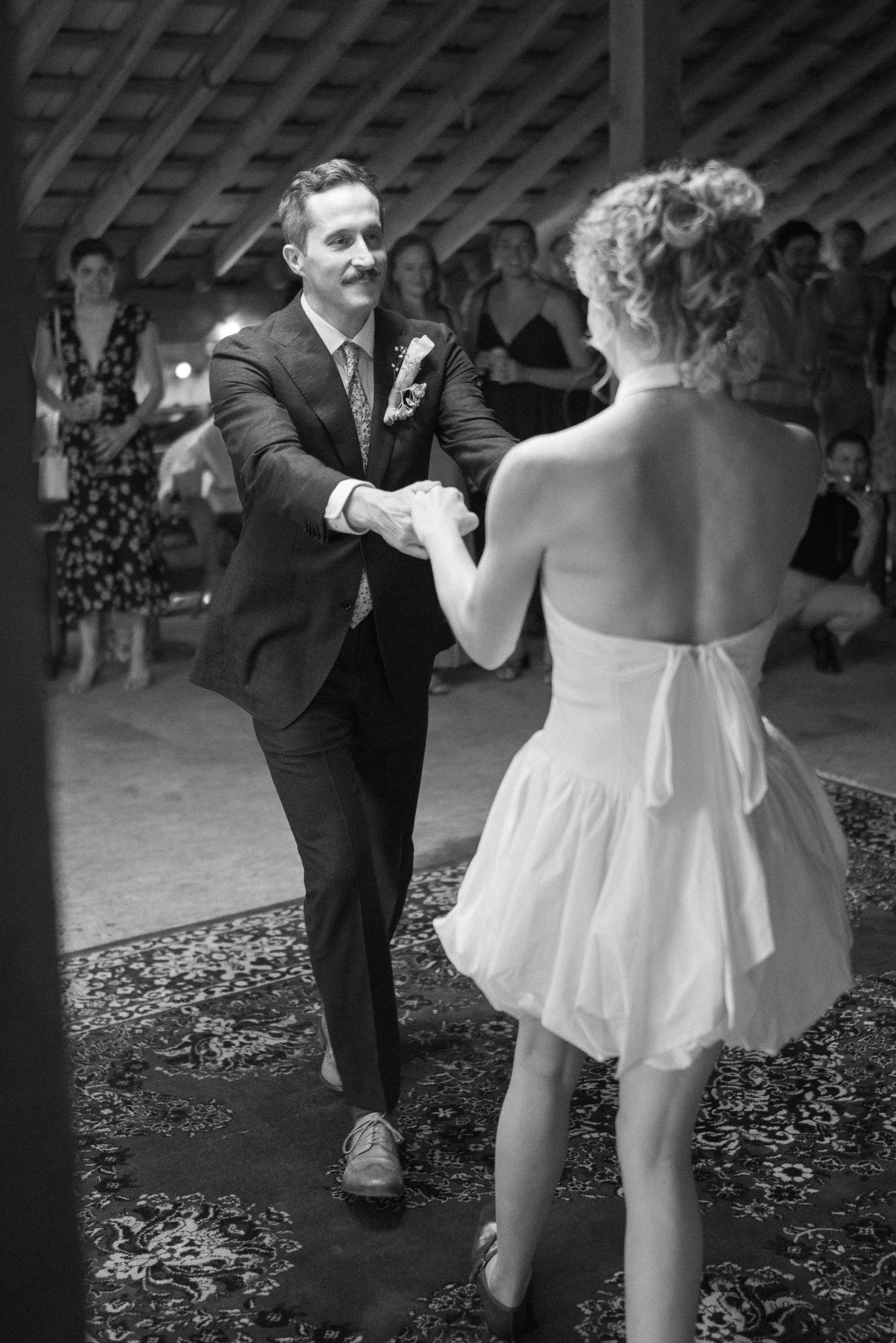 A black and white photo of a wedding reception where a man in a suit is dancing with a woman in a dress. Other guests are watching in the background.