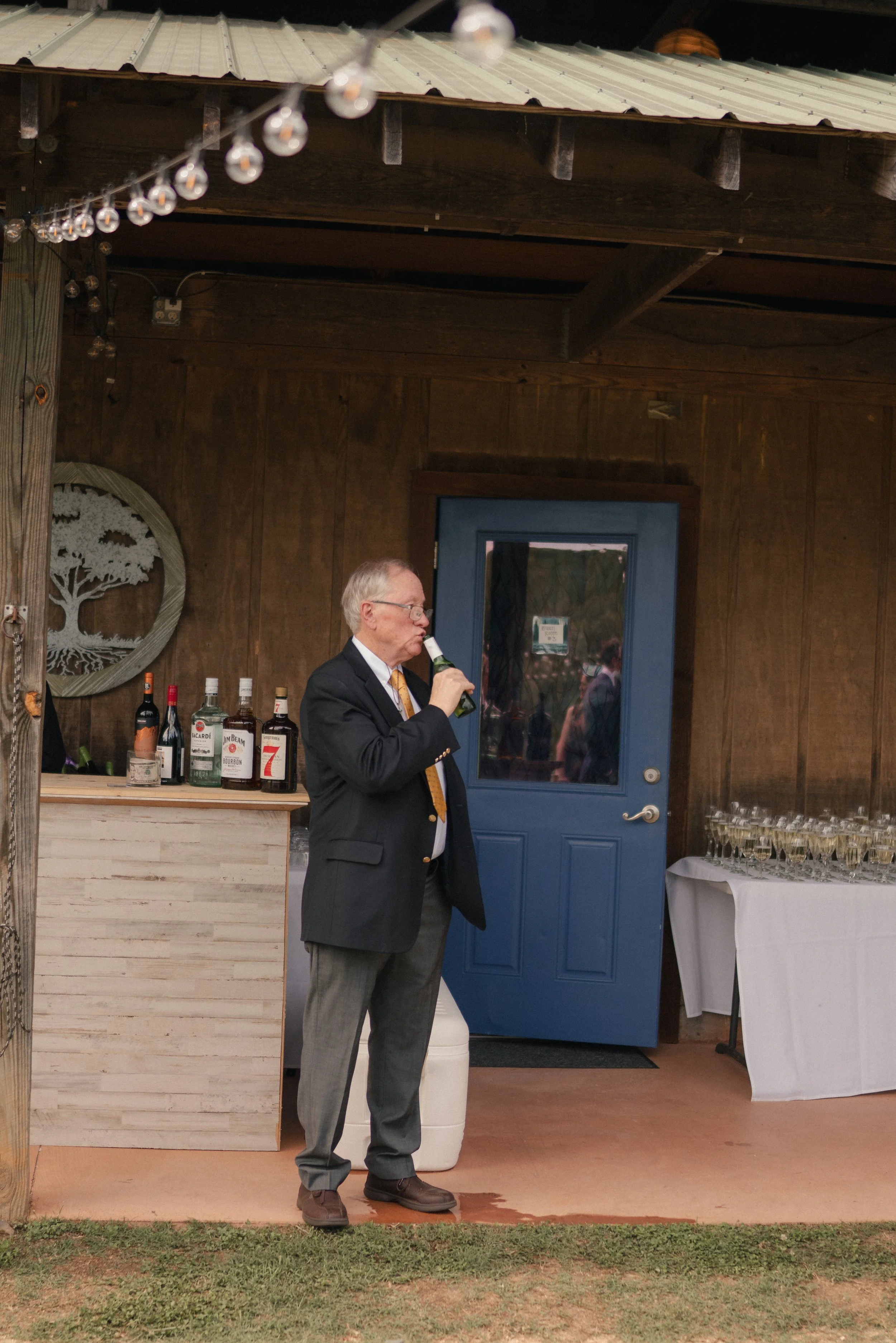 A man in a suit and tie, with gray hair and glasses, is drinking from a bottle at a bar area with various alcohol bottles behind him. The setting appears to be indoors with wooden walls, string lights overhead, and a blue door in the background. To t