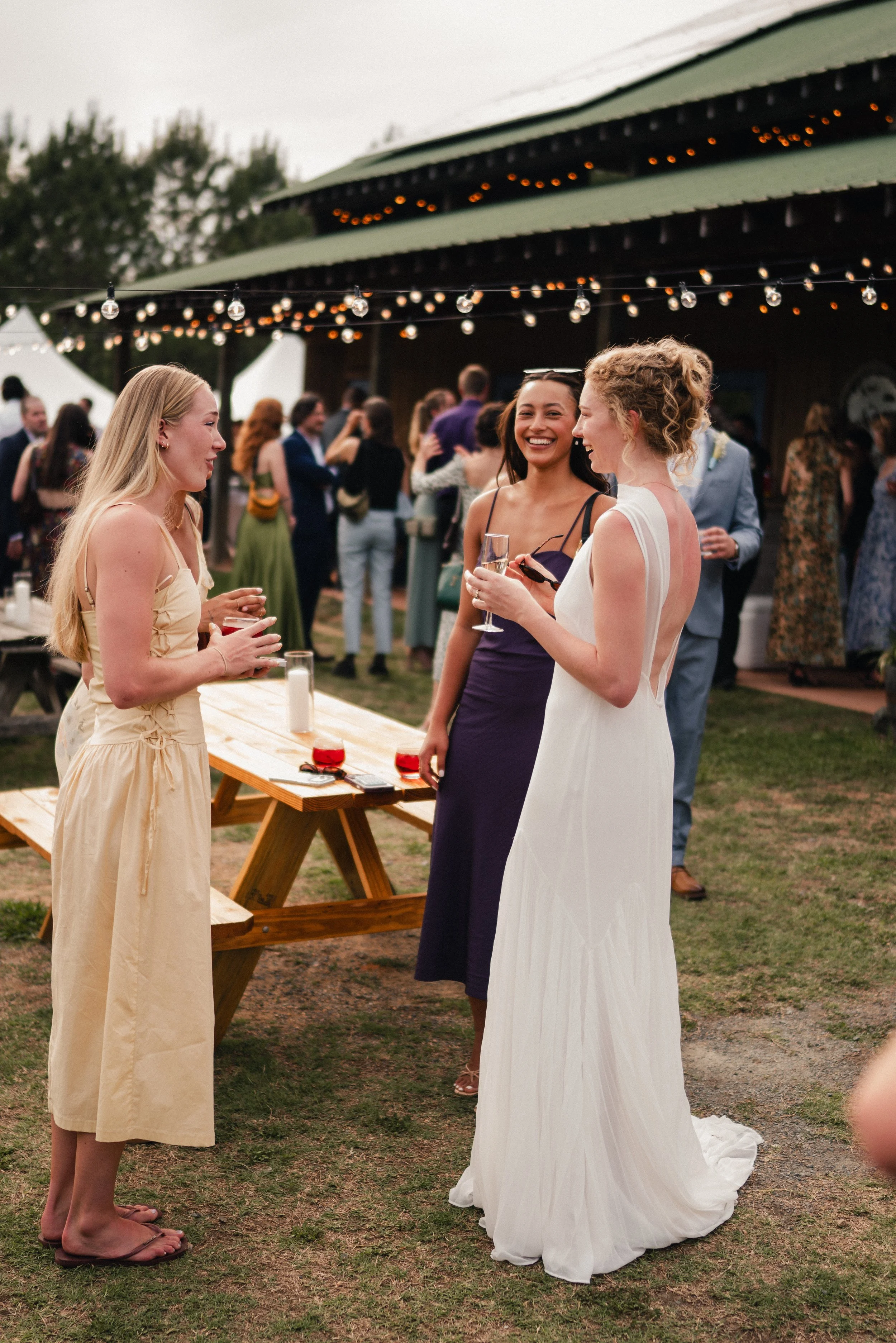 Three women in formal attire, two in dresses and one in a gown, standing and conversing outdoors at an event with a crowd in the background, string lights overhead, and a green roofed structure nearby.