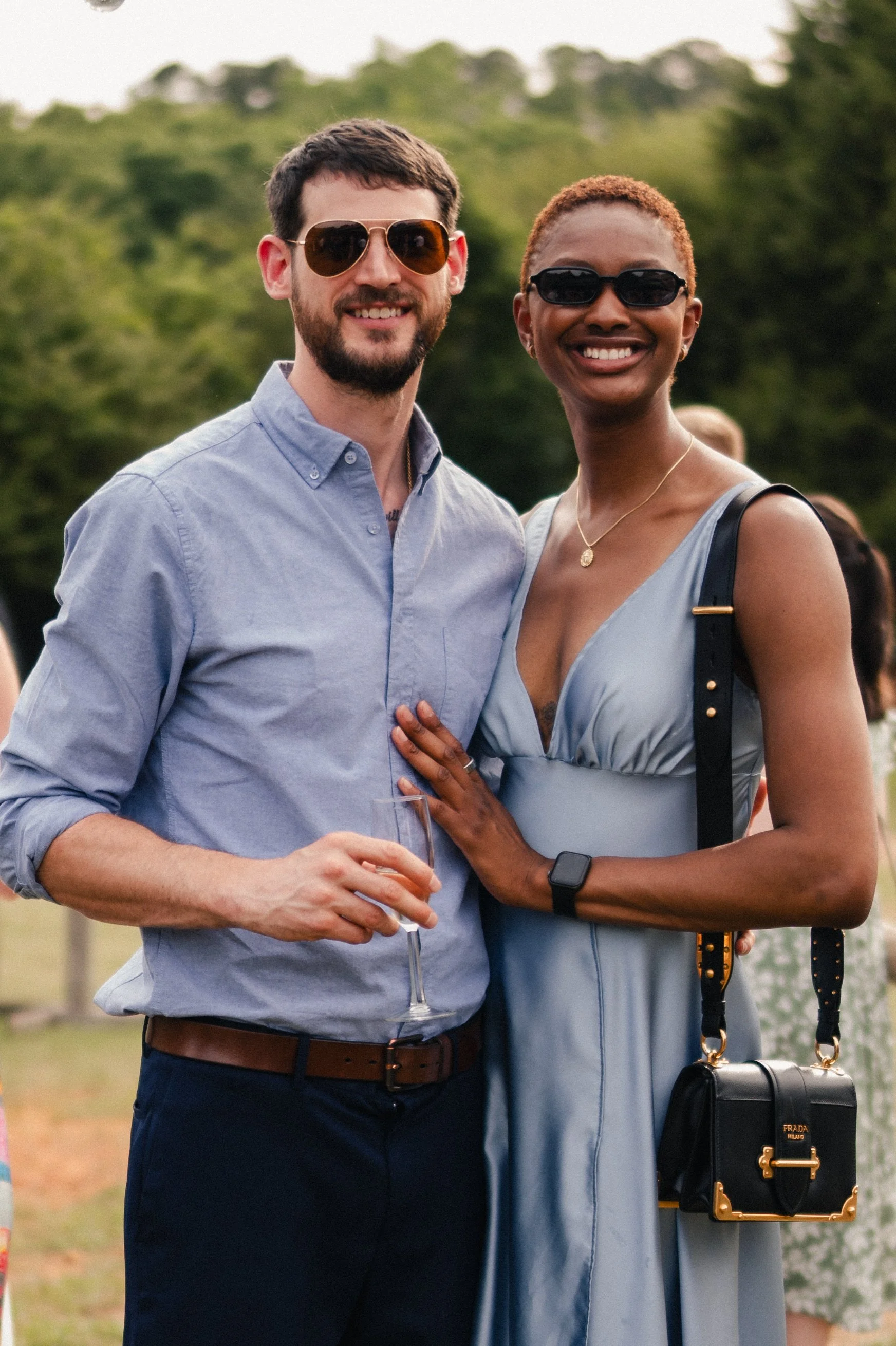 A man with a beard and glasses holding a wine glass, and a woman with short hair and glasses smiling, standing close together outdoors with trees in the background.