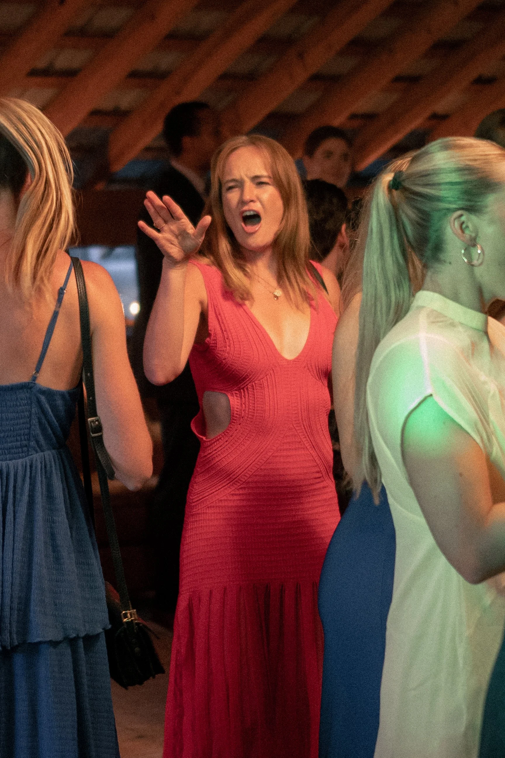 A woman with red hair in a red dress appears to be angry or shouting, with her mouth open and hand raised, in a social setting with other women and men around her.