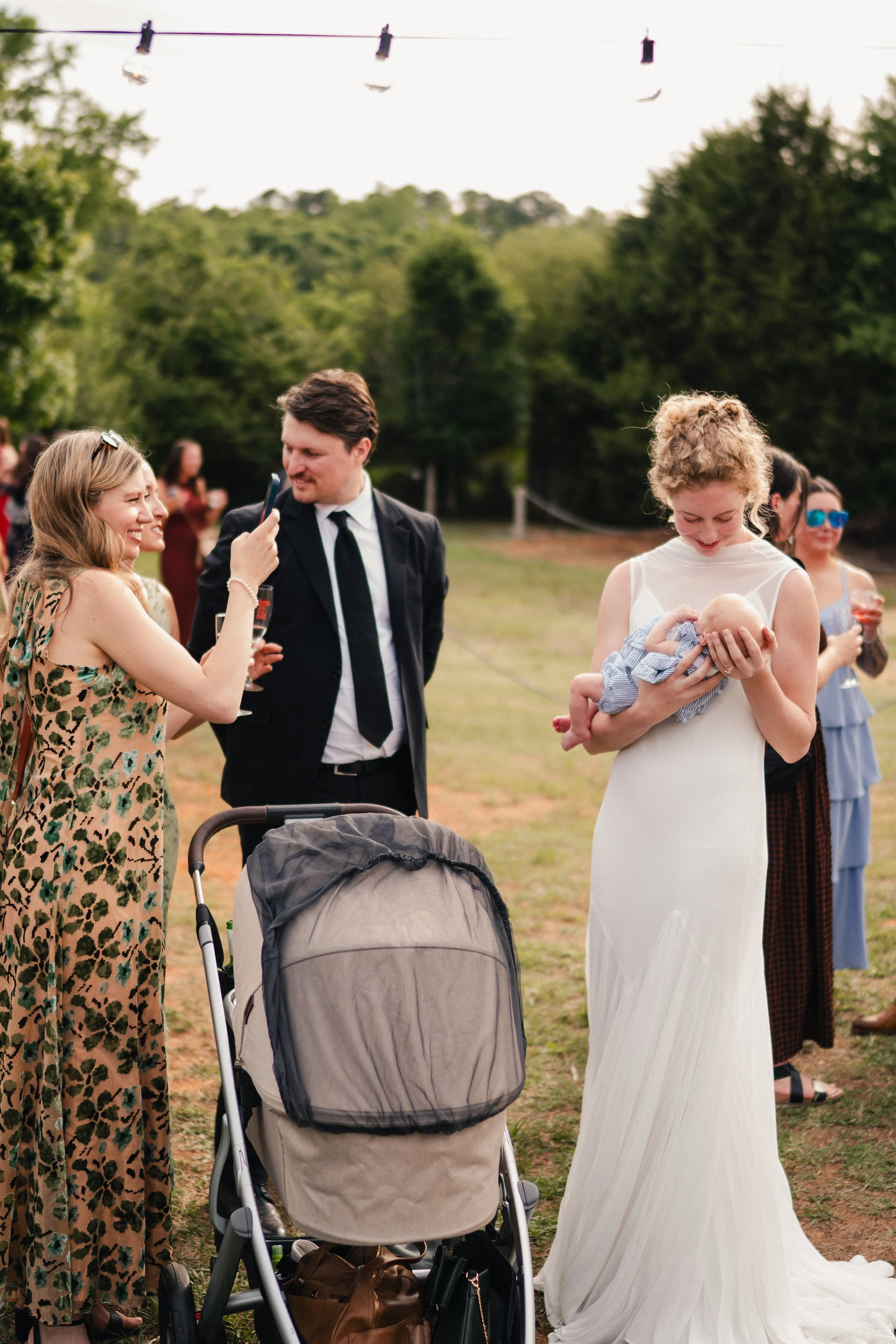 A group of people at an outdoor event, including a woman in a white dress holding a baby, a woman in a leopard-print dress, and a man in a suit, with trees and string lights in the background.