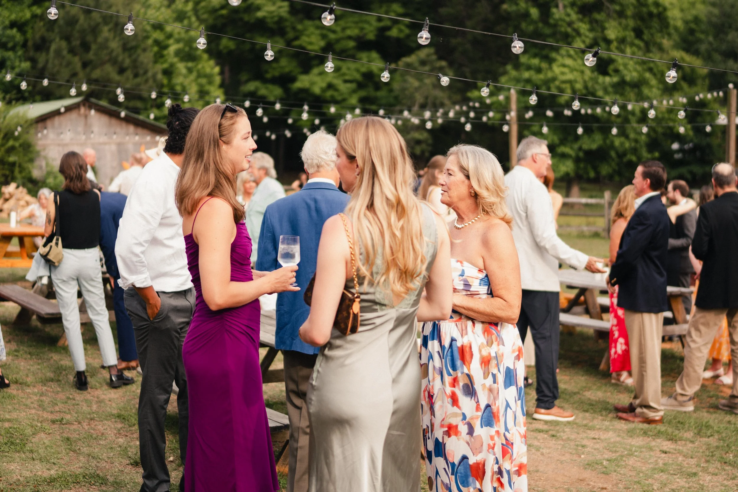A group of people socializing outdoors at a gathering, with string lights overhead and trees in the background.