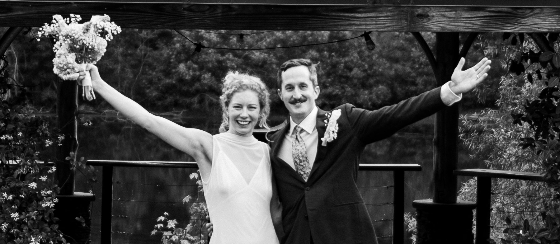 Black and white photograph of a happy bride and groom celebrating outdoors, with arms raised and smiles on their faces. The bride is holding a bouquet of flowers, and the groom is dressed in a suit with a floral tie and boutonniere. They are standing