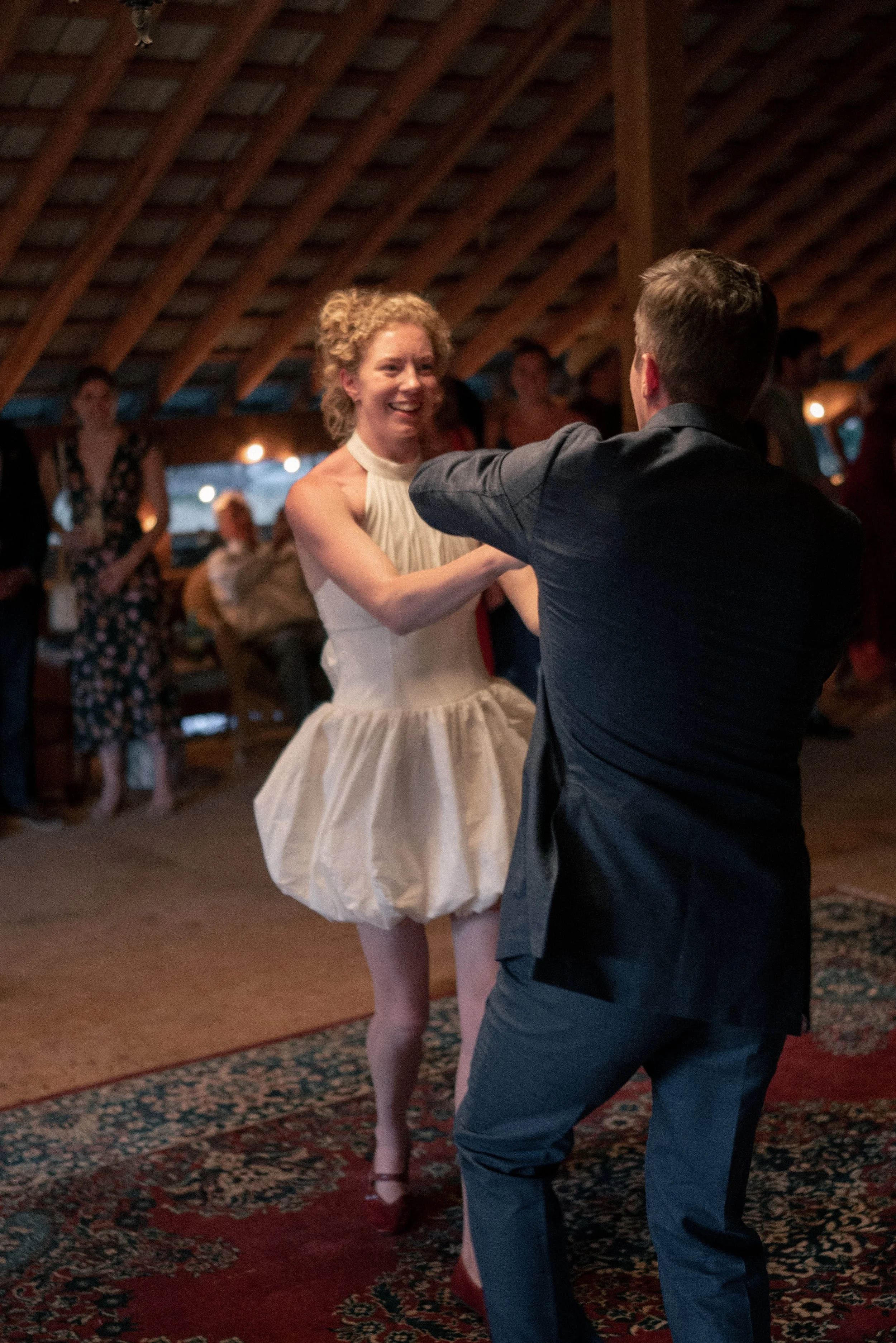 A woman with curly blonde hair in a white dress is dancing with a man in a dark suit. They are in a warmly lit indoor setting with a wooden, curved ceiling. Other people are visible in the background, watching and mingling.