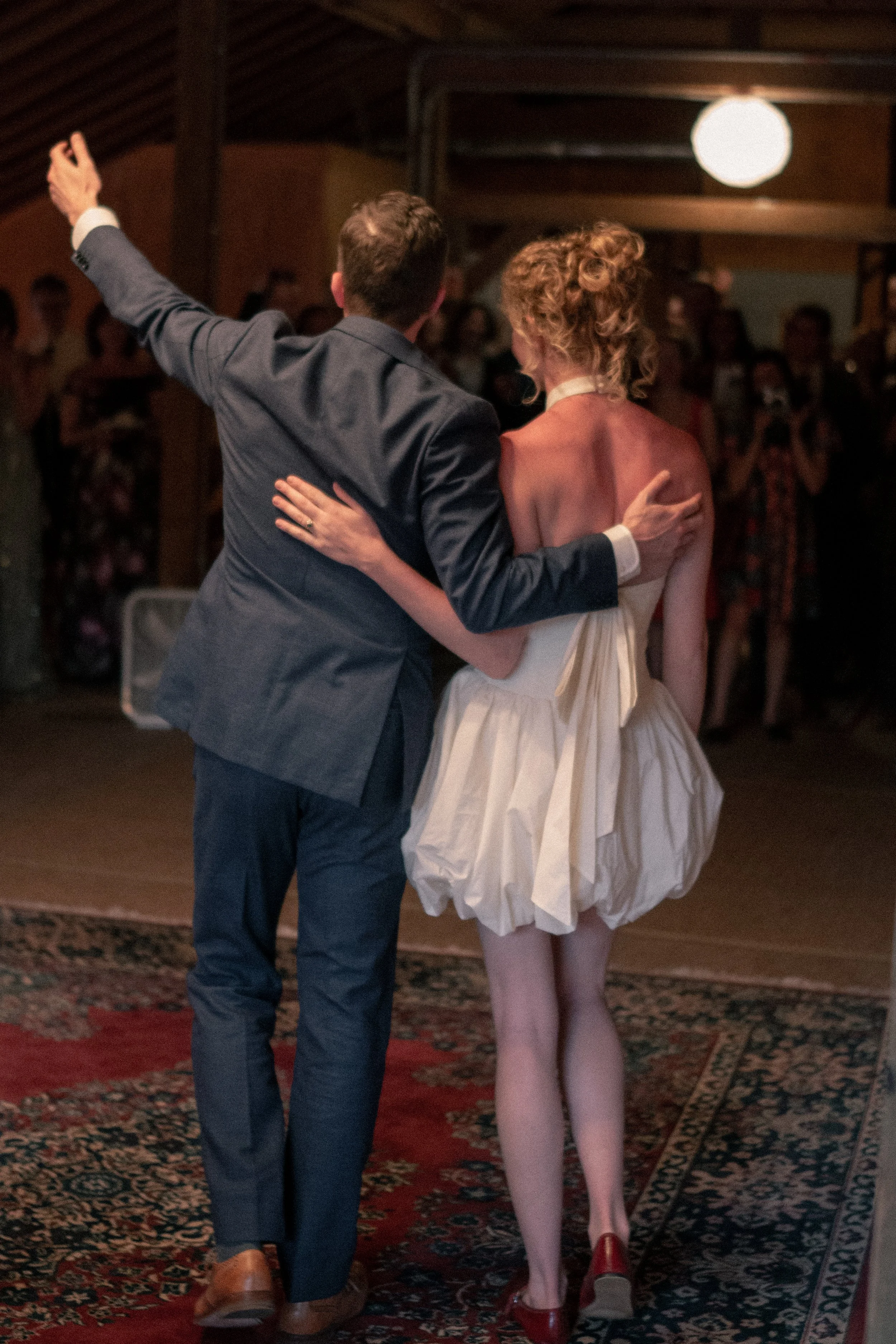 A man and woman dancing closely in a dimly lit room filled with people, with the man raising one arm and the woman wearing a strapless dress and red shoes.