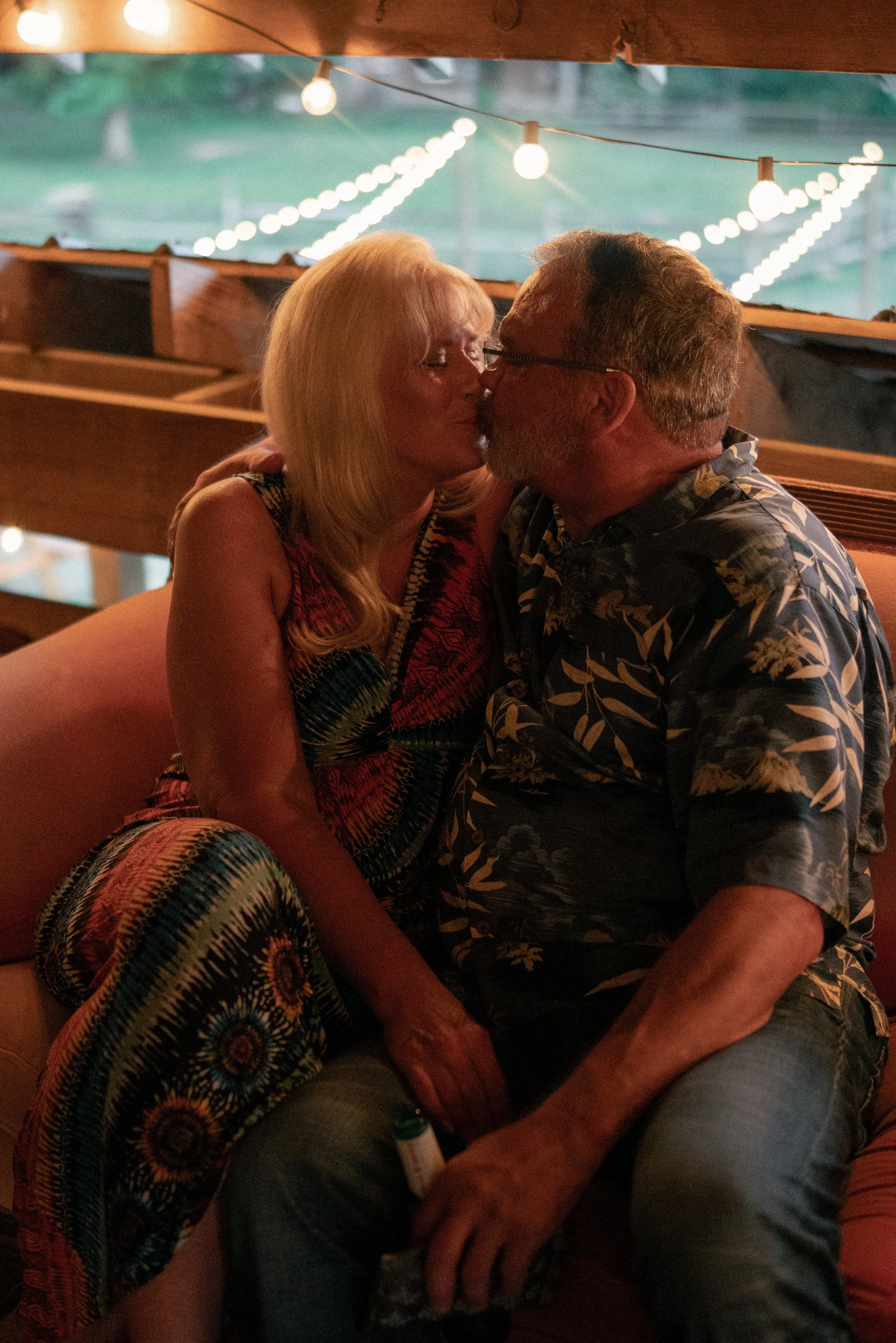 A couple sharing a kiss at a nighttime outdoor setting decorated with string lights.