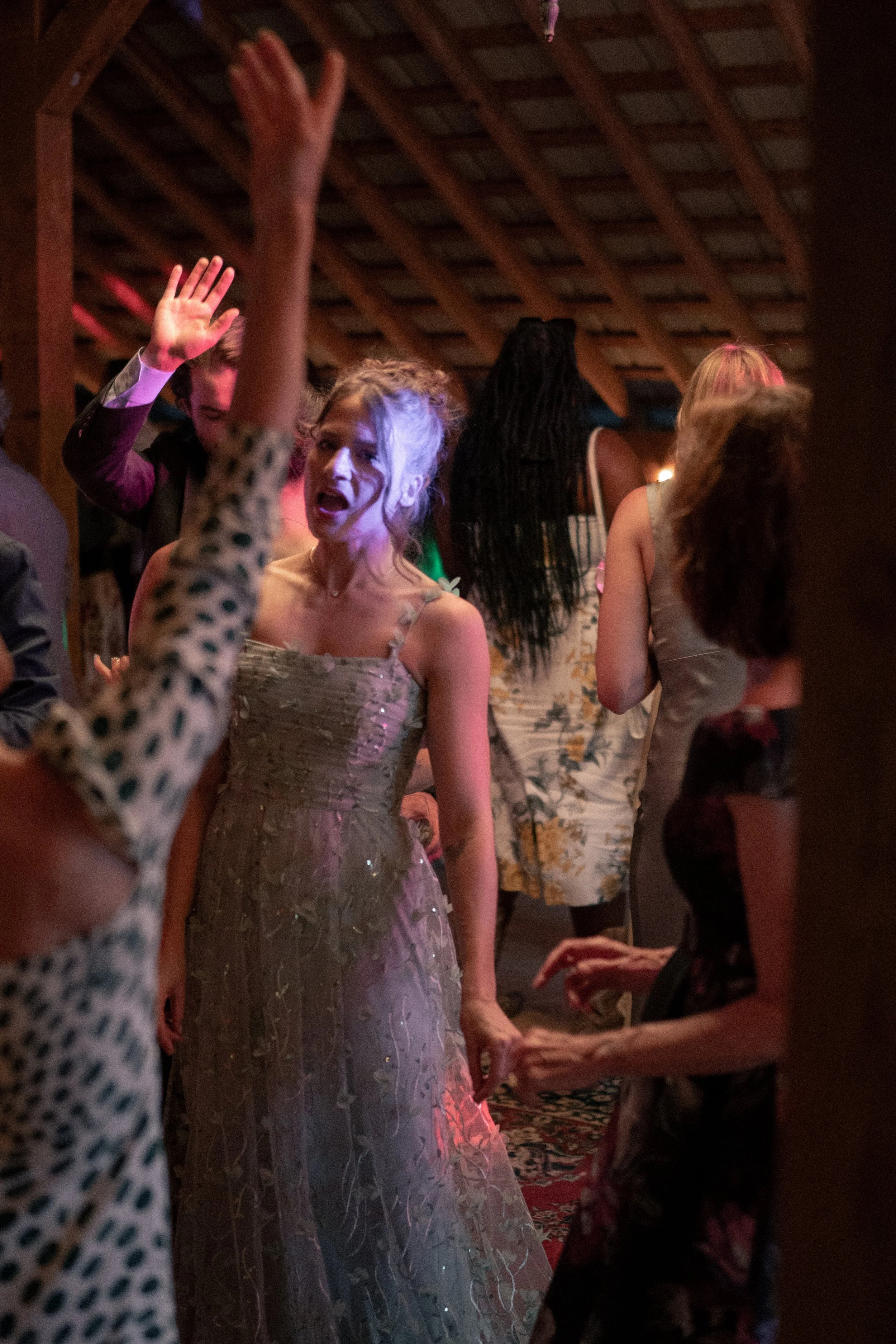 People gathered at a party, with one woman in a pink and silver dress raising her hand and speaking, others are socializing under a wooden ceiling with warm lighting.