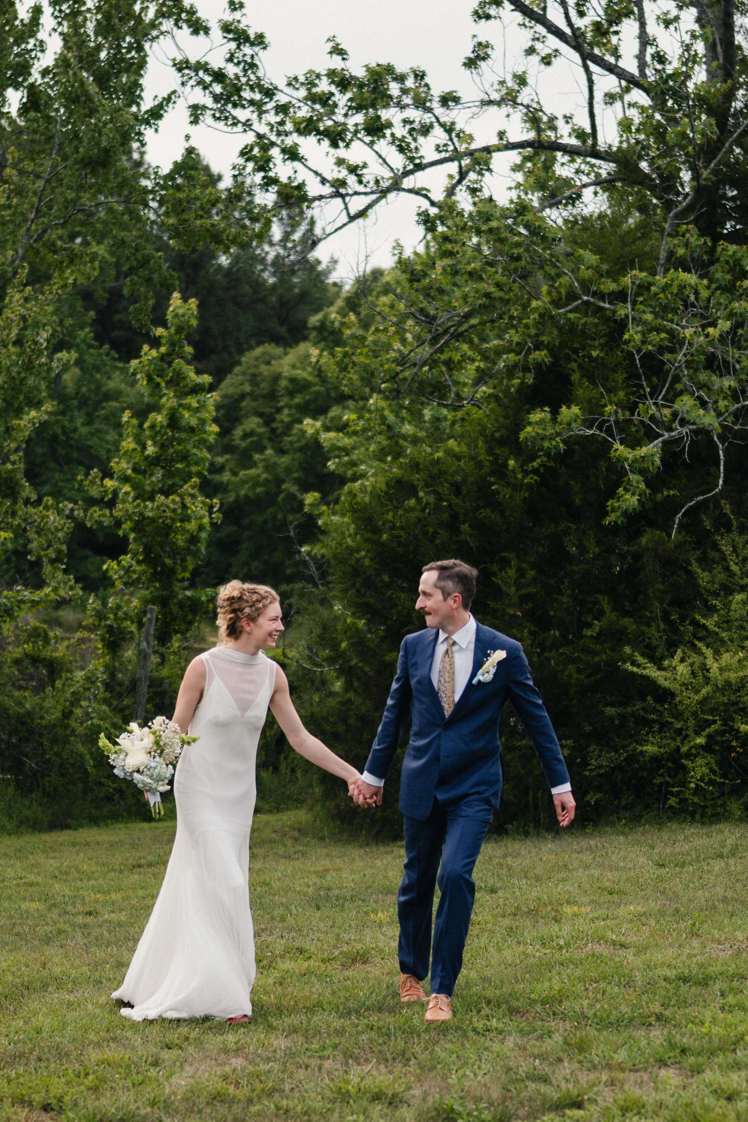A bride and groom walking hand in hand outdoors on a grassy lawn, with trees in the background, during their wedding celebration.