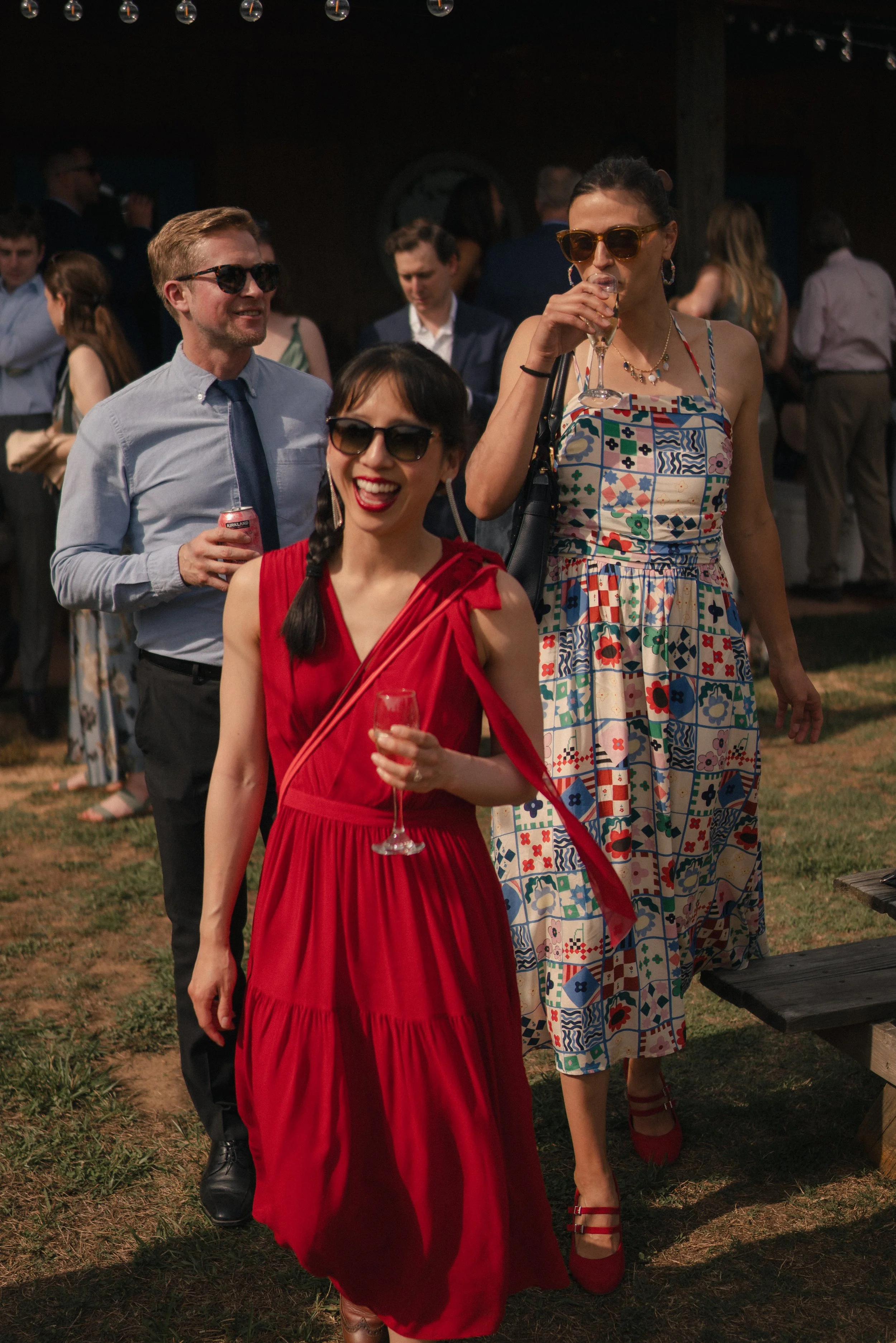 People at a social event, with women in colorful dresses and men in suits, holding drinks and smiling outdoors in the evening.