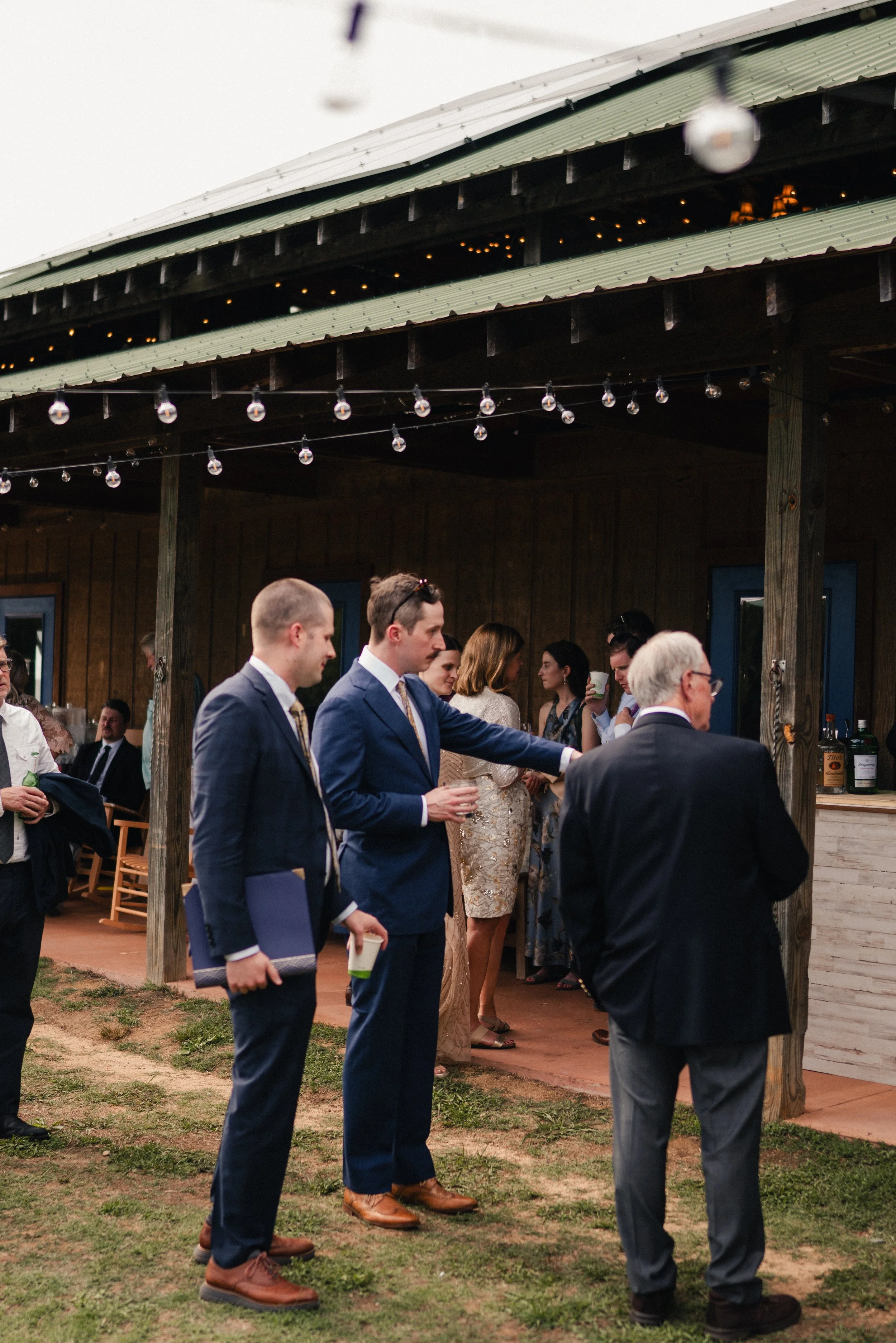 Group of people dressed in formal attire at a social gathering outdoors, standing under a wooden structure with string lights. Some individuals are holding drinks and engaging in conversation.