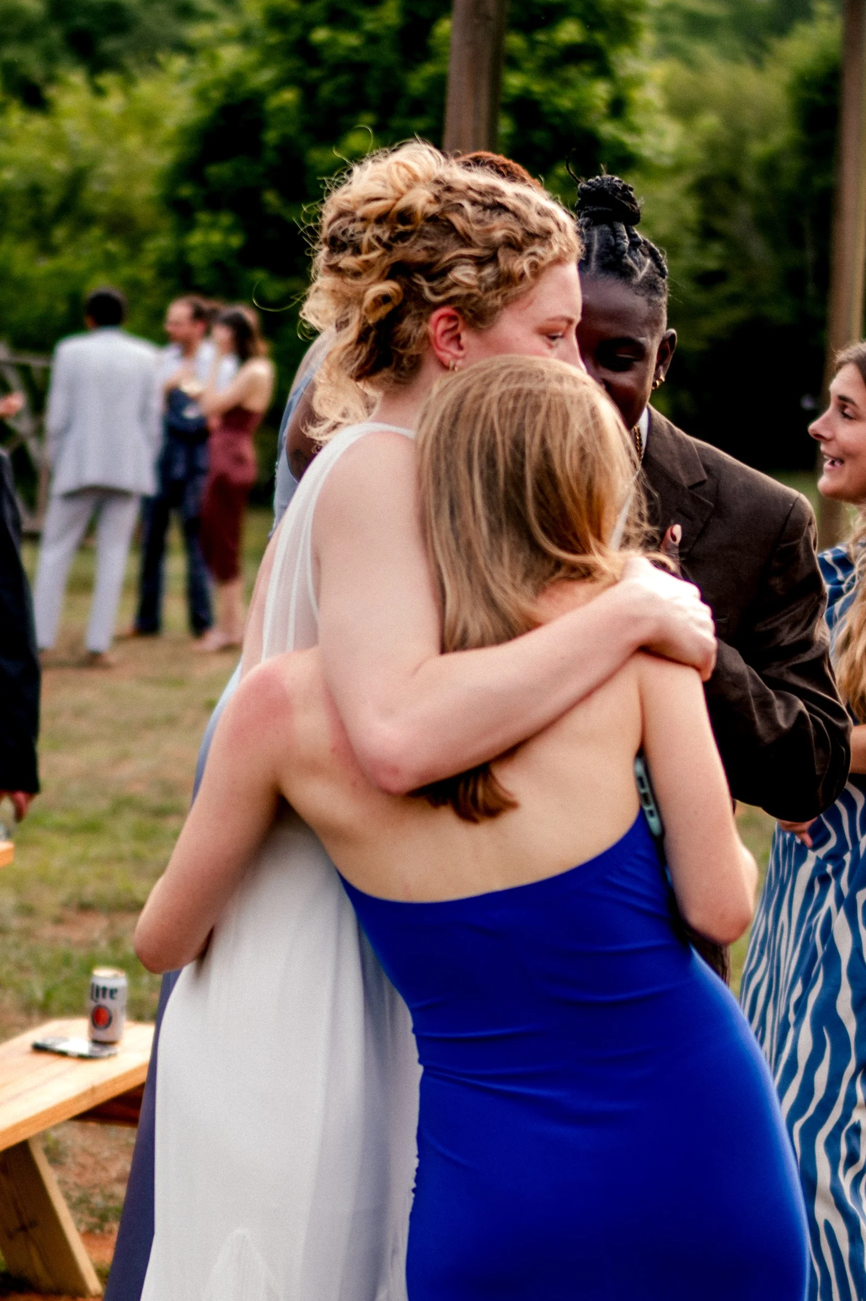 People hugging at an outdoor gathering, with a woman in a white dress and another in a blue dress in the foreground, and blurred background of more people and trees.