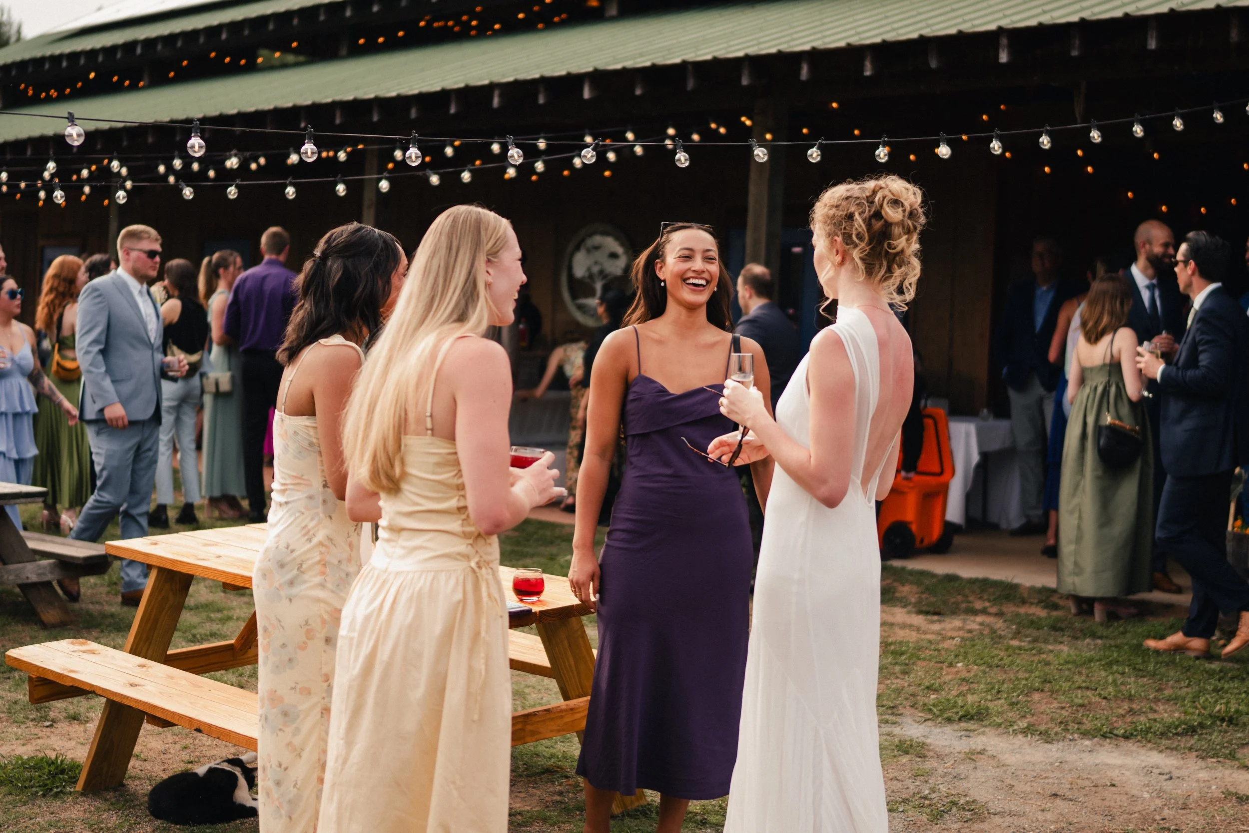 A group of women are socializing at an outdoor event, standing and talking near a picnic table with drinks in their hands. People are dressed in summer attire, and there are string lights hanging above. A barn-like building is in the background.