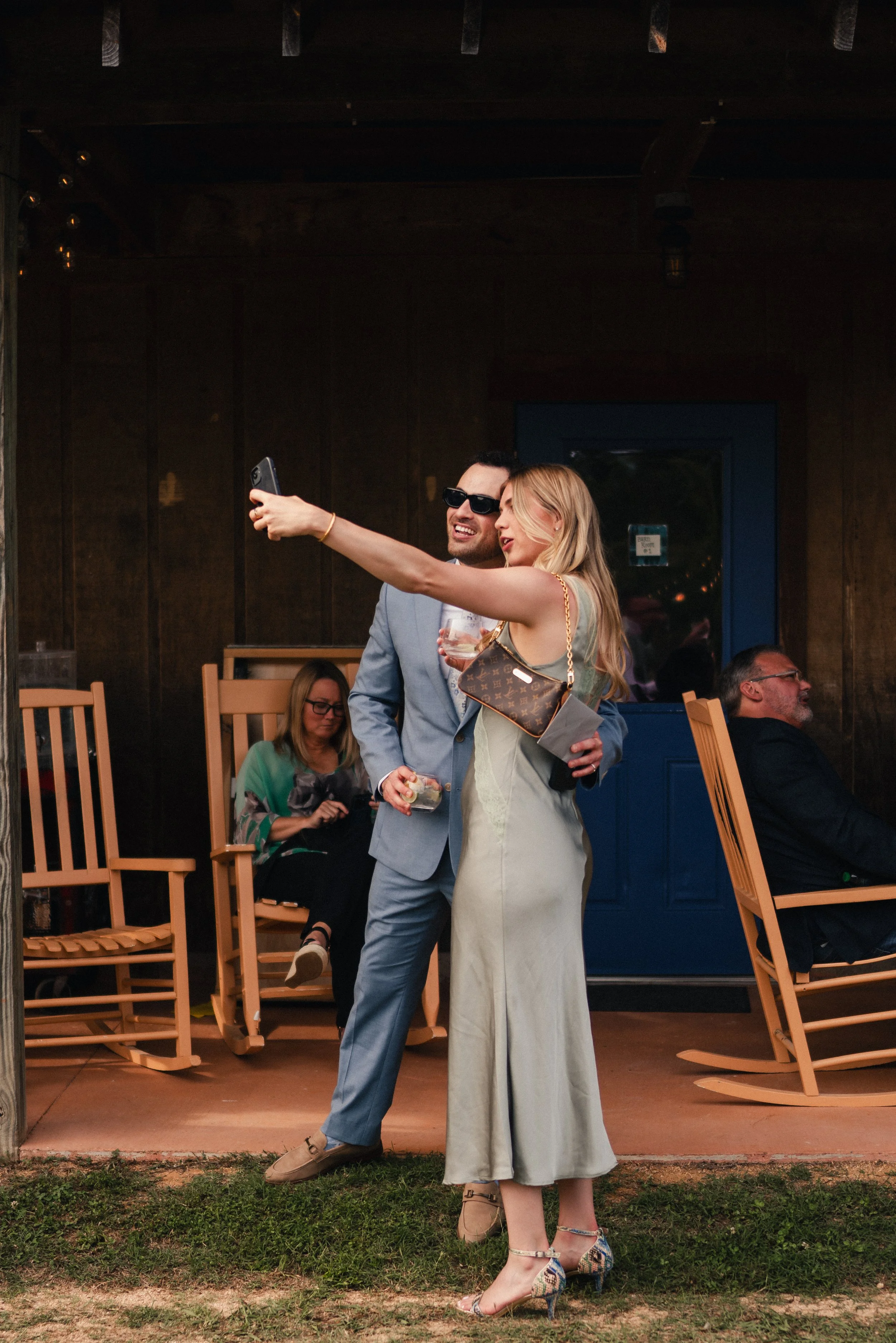A man in a light blue suit and sunglasses taking a selfie with a woman in an off-the-shoulder dress, holding a wine glass, at an outdoor event. Multiple seated people are in the background, some looking at the camera, with a wooden wall and string li