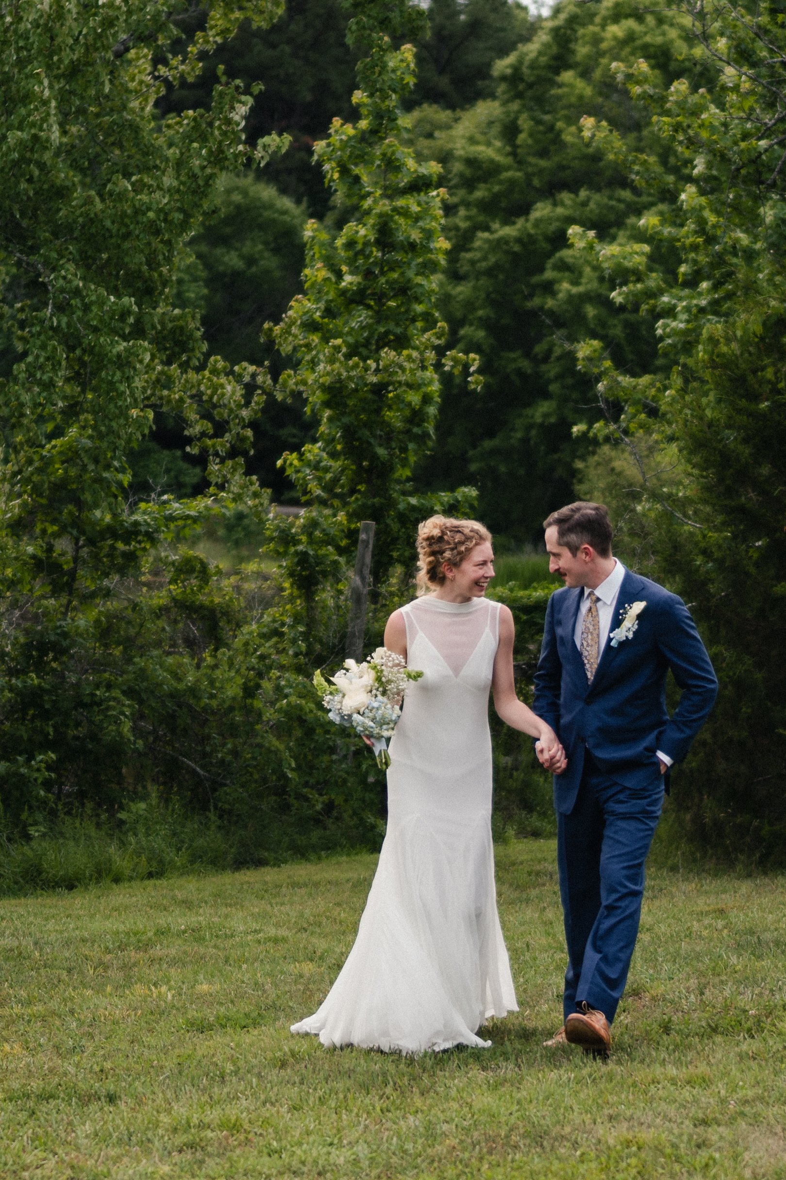 A bride and groom holding hands and smiling at each other, outdoors on a grassy lawn with trees in the background.