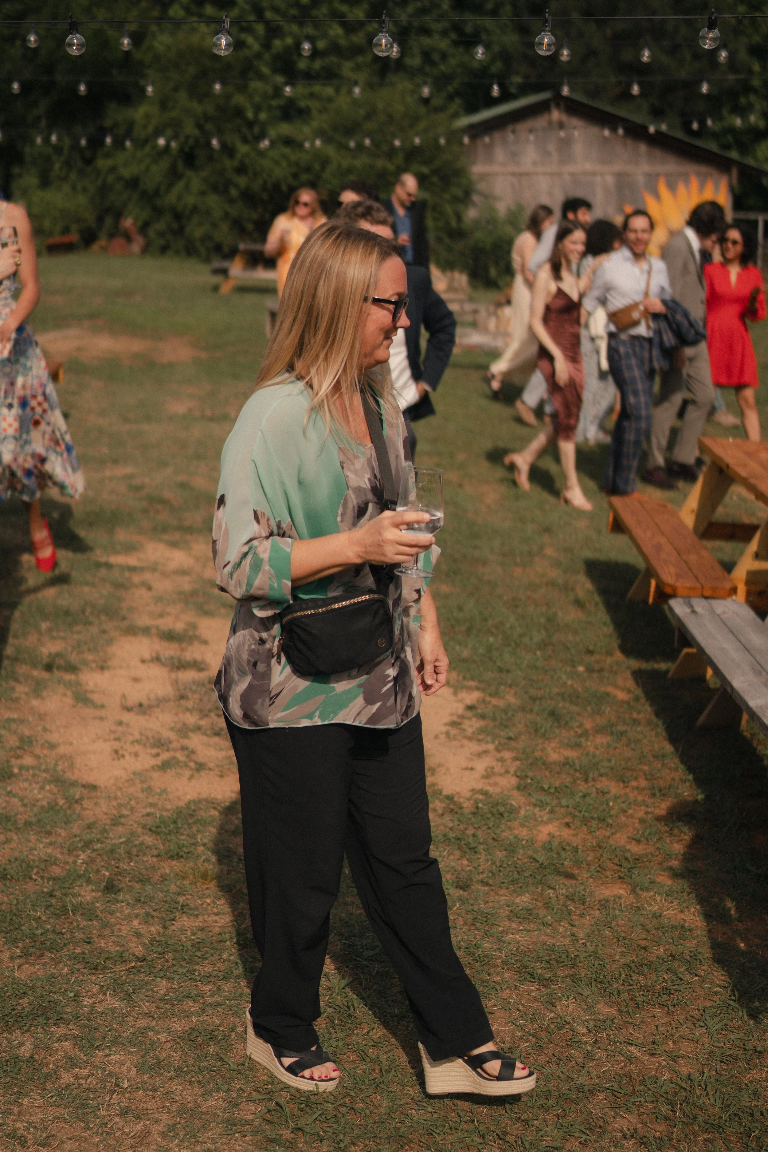 A woman with blonde hair and sunglasses holding a glass of water walking outdoors at an evening gathering with string lights, other people socializing, and a wooden building in the background.