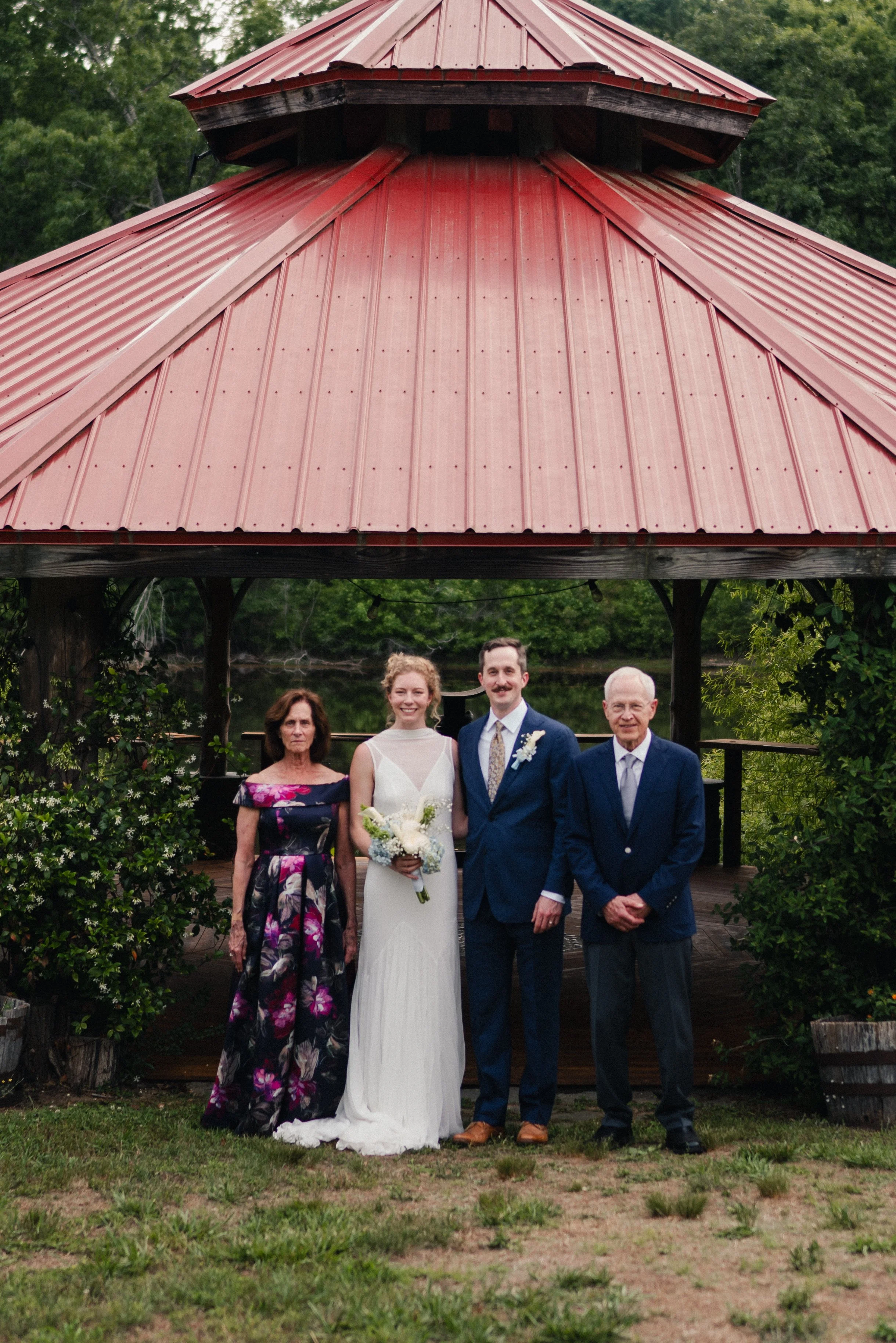 Group of five people, including a bride and groom, standing outdoors in front of a wooden gazebo with a red metal roof, surrounded by greenery.