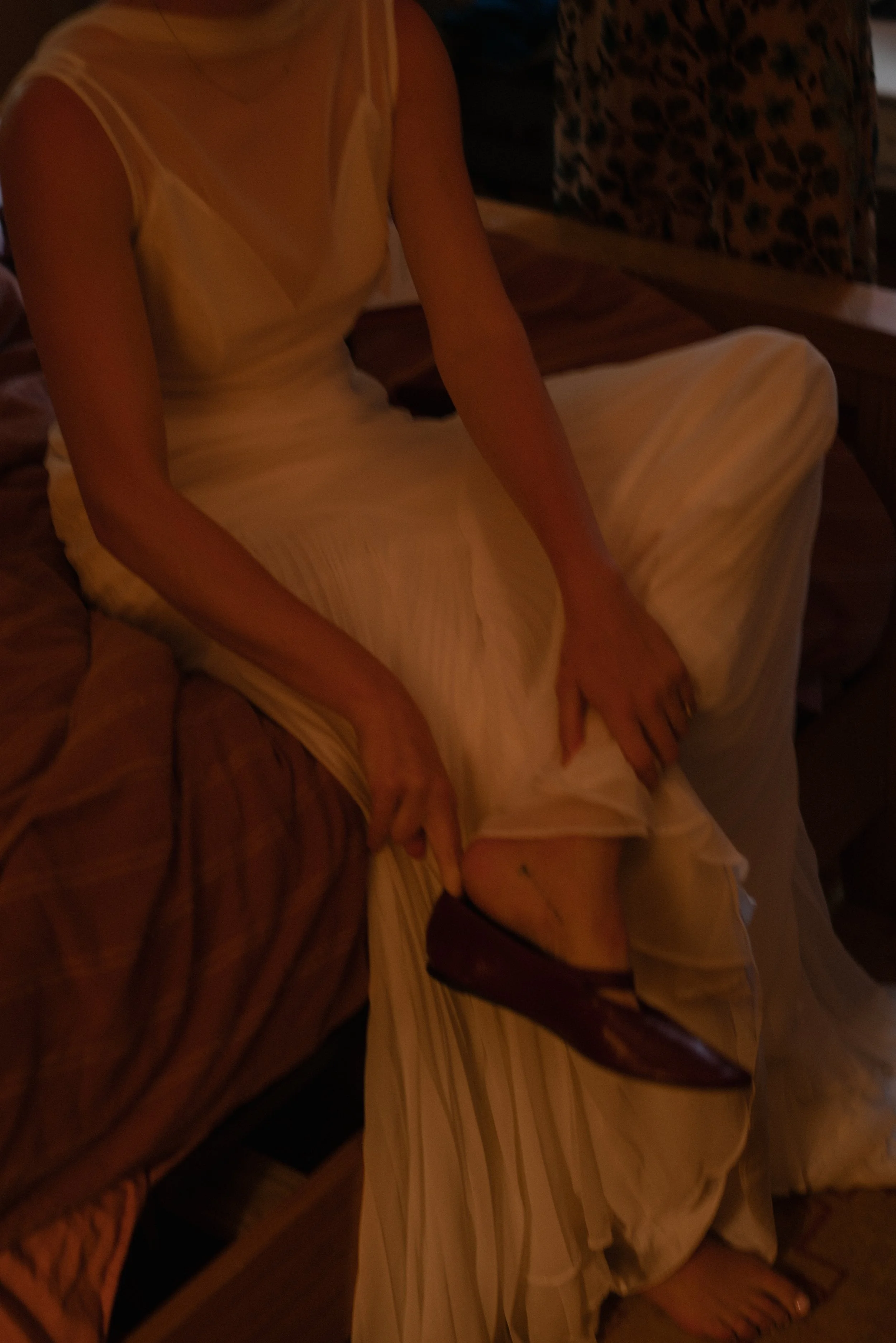 A woman seated on a dark wooden chair, wearing a sleeveless, light-colored dress with a sheer top and a flowing skirt, with her hands resting on her lap and her right foot slightly visible.