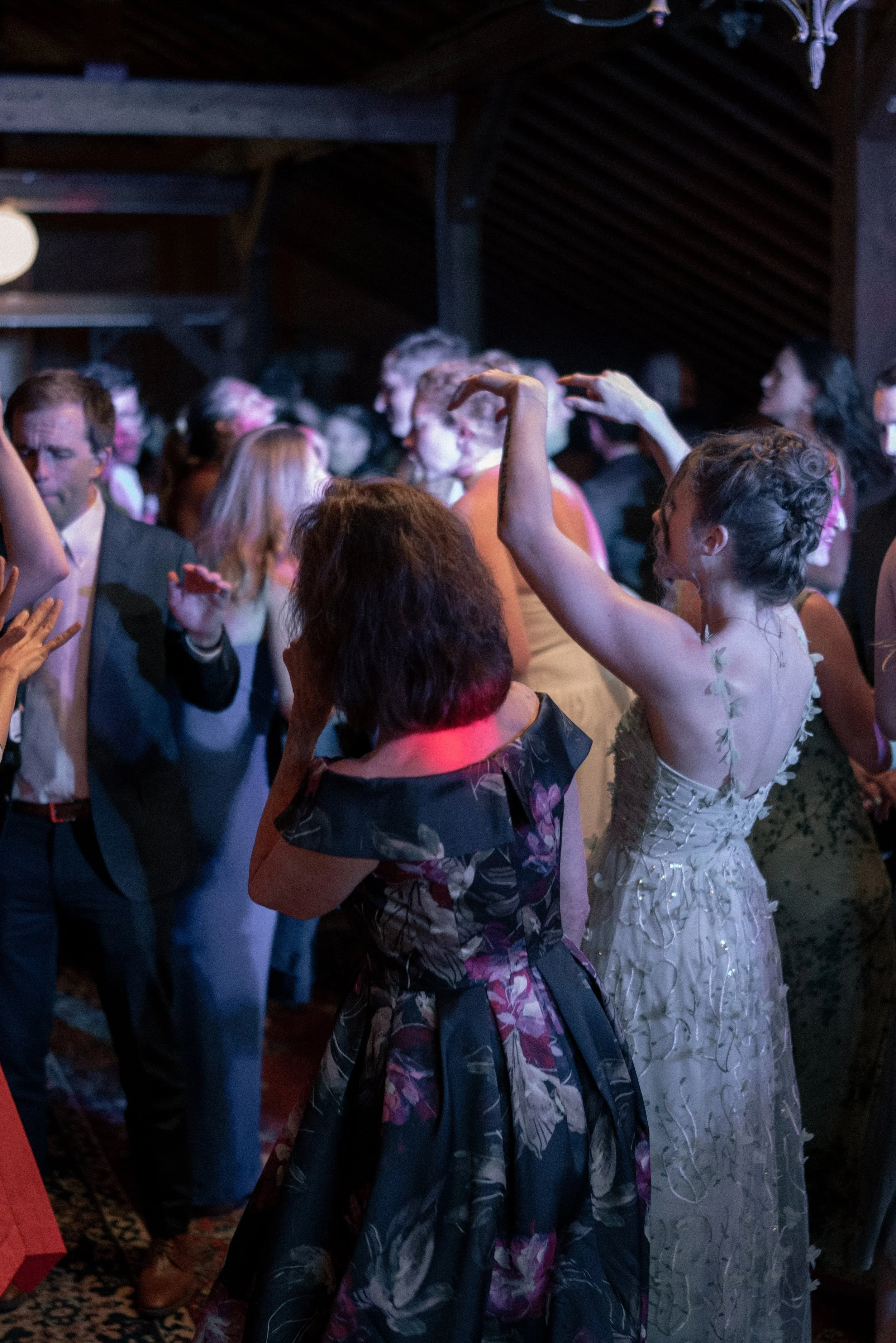 A group of people dancing at a party or celebration, with women in floral dresses and men in suits, under dim lighting in an indoor venue.
