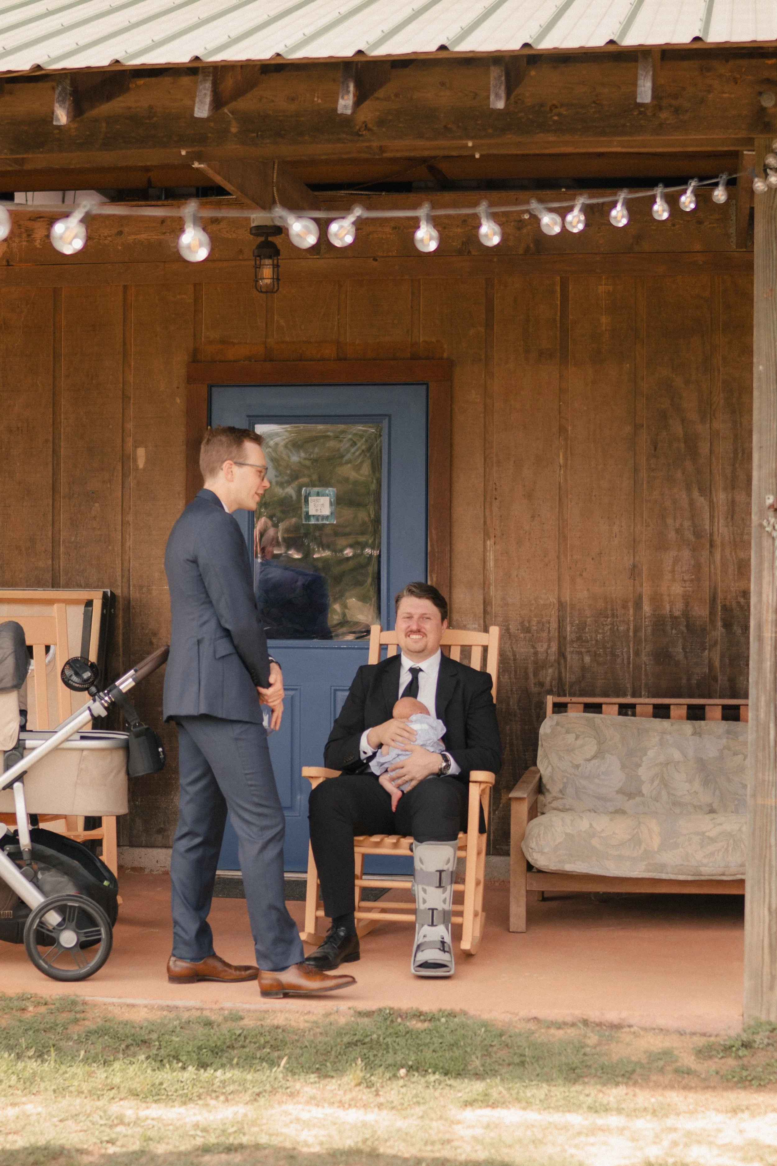 A man in a suit with a leg brace and crutches sitting on a wooden chair, holding a baby; another man in a suit standing beside him, smiling and looking down at the baby; outdoor setting with wooden walls, string lights, and a blue door in the backgro