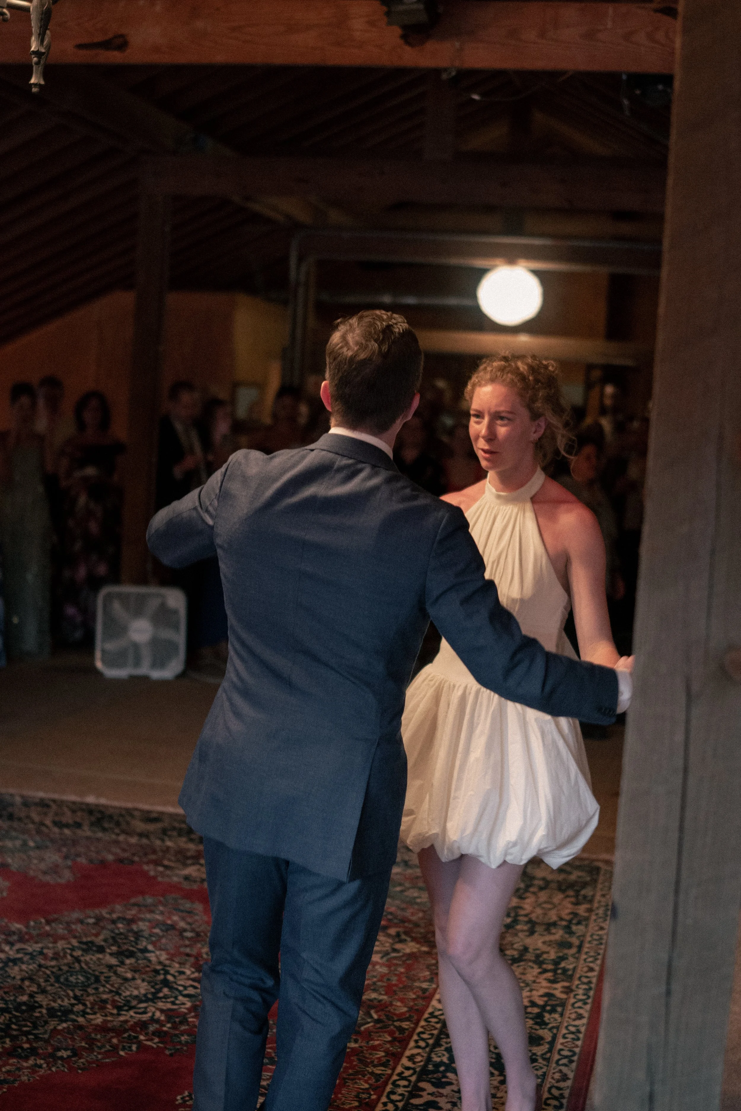A man in a gray suit dancing with a woman in a cream-colored dress at a social gathering indoors. The woman appears to be surprised or emotional while dancing. Other people are visible in the background, watching or waiting for their turn to dance. T