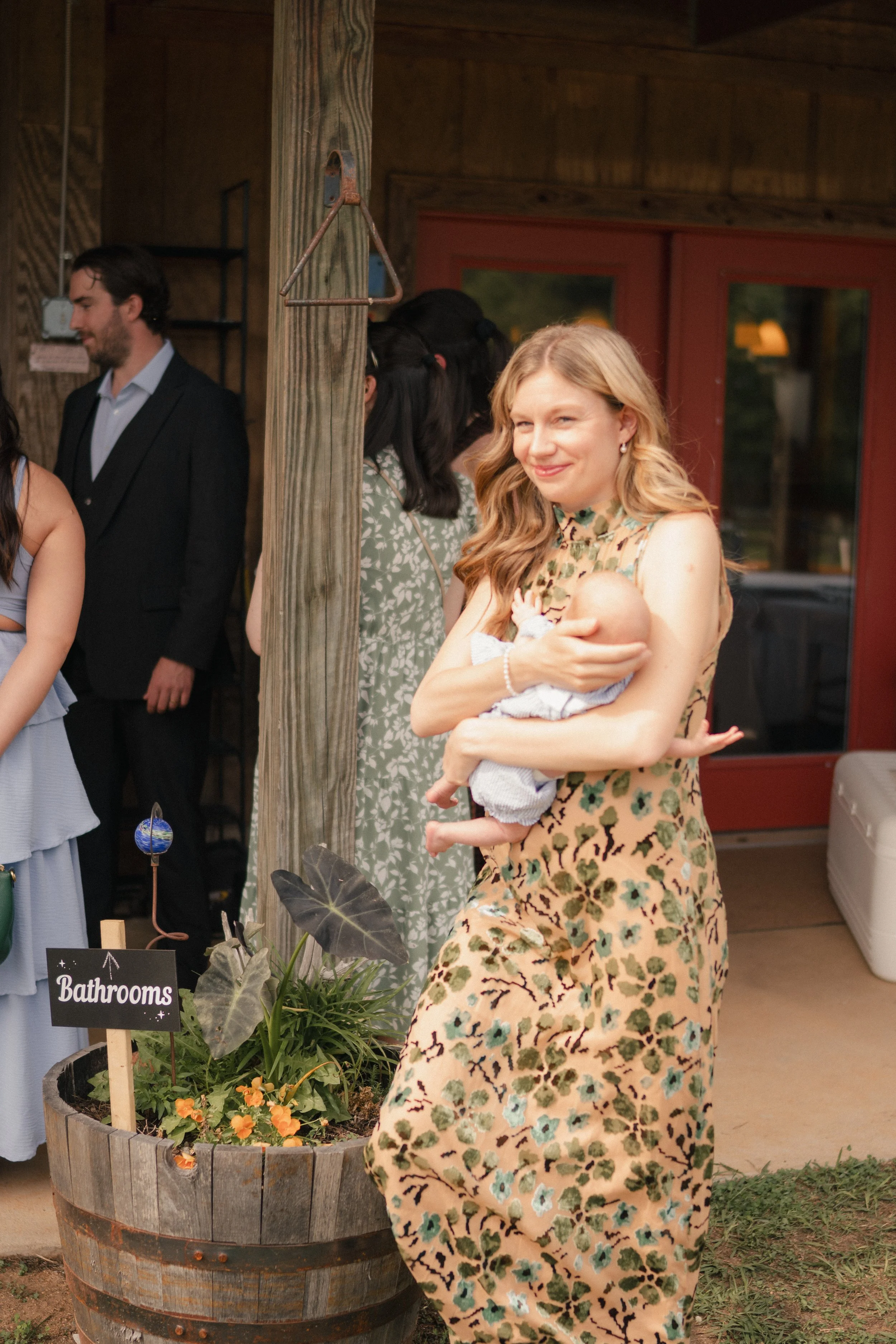 A woman holding a baby, standing outdoors near a wooden flower planter with orange flowers, at an event marked by a sign that says 'Bathrooms'.