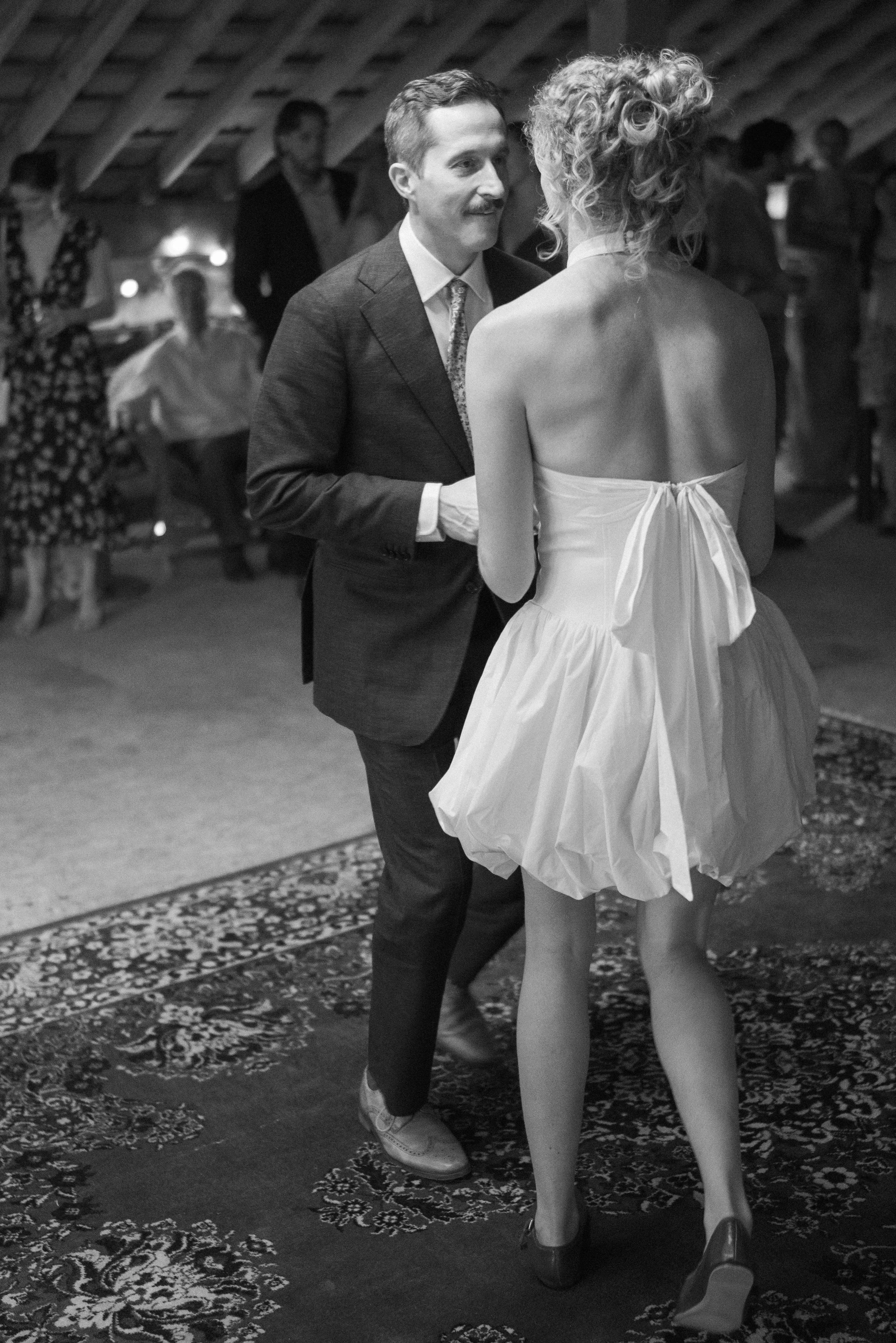 A man and woman are dancing closely at a social event in an indoor venue with a patterned carpet and wooden ceiling beams. The man is wearing a suit and tie, and the woman is wearing a strapless dress and high heels. Other people are visible in the b