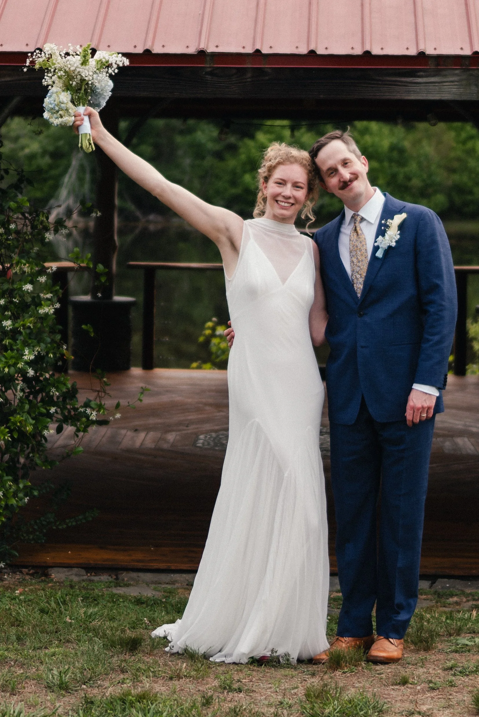 A smiling woman in a white wedding dress holding a bouquet raises her arm while standing next to a man in a blue suit for a wedding photoshoot outdoors near a wooden pavilion with a red metal roof, surrounded by greenery.