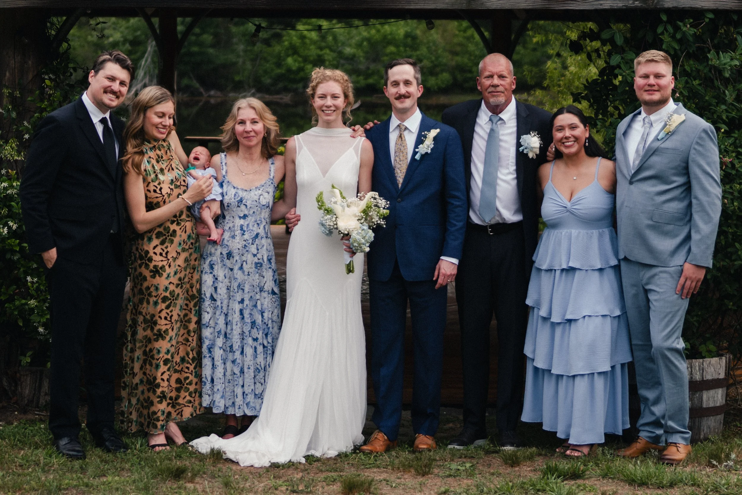 A diverse group of nine people, including a bride and groom, posing outdoors at a wedding ceremony. The bride wears a white dress and holds a bouquet; the groom wears a blue suit with a boutonniere. The others are dressed in formal attire, some in dr