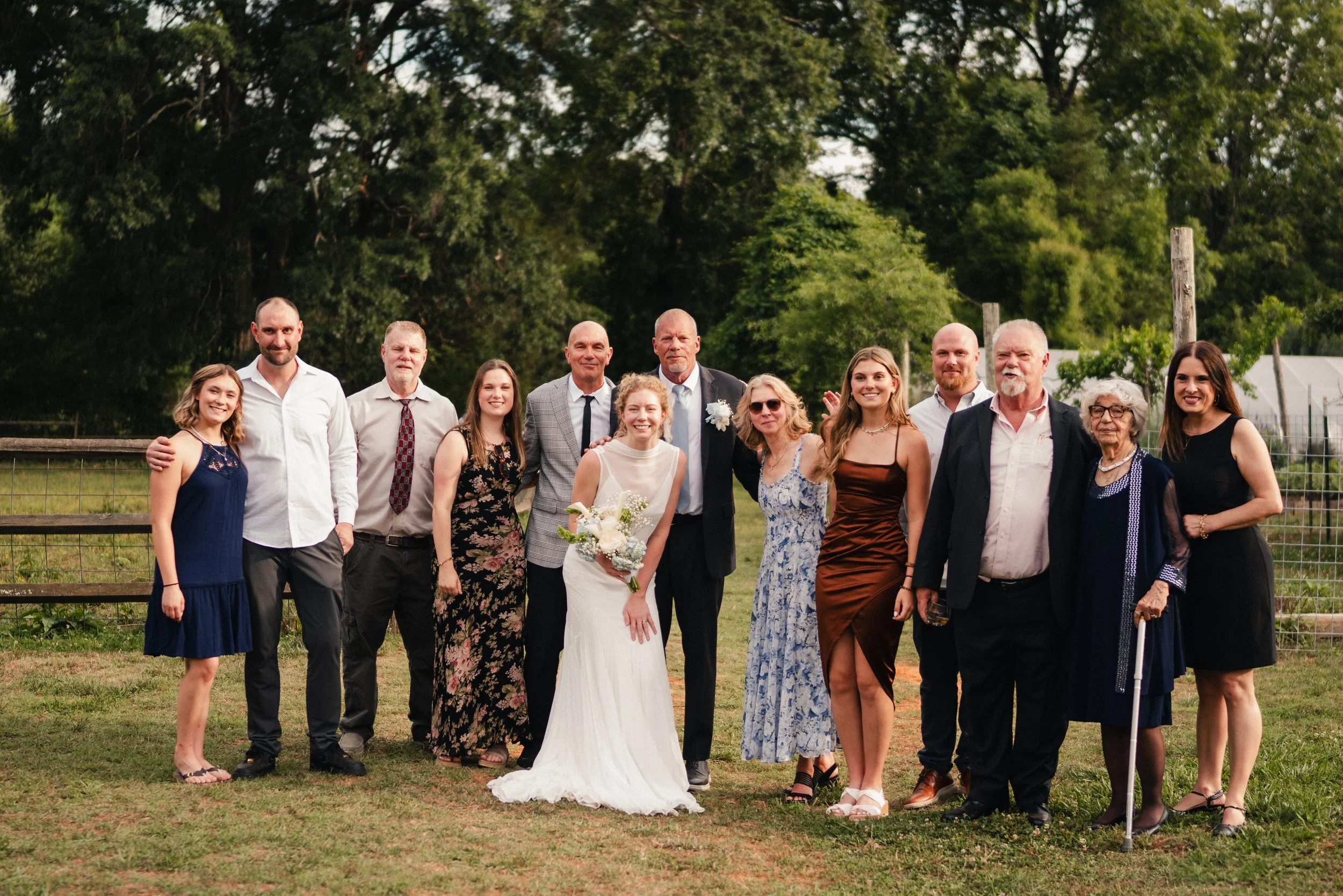 A group of people, including a bride in a white dress holding a bouquet and a groom in a gray plaid suit, standing outdoors with a backdrop of green trees. They are smiling and dressed in formal attire, likely celebrating a wedding.