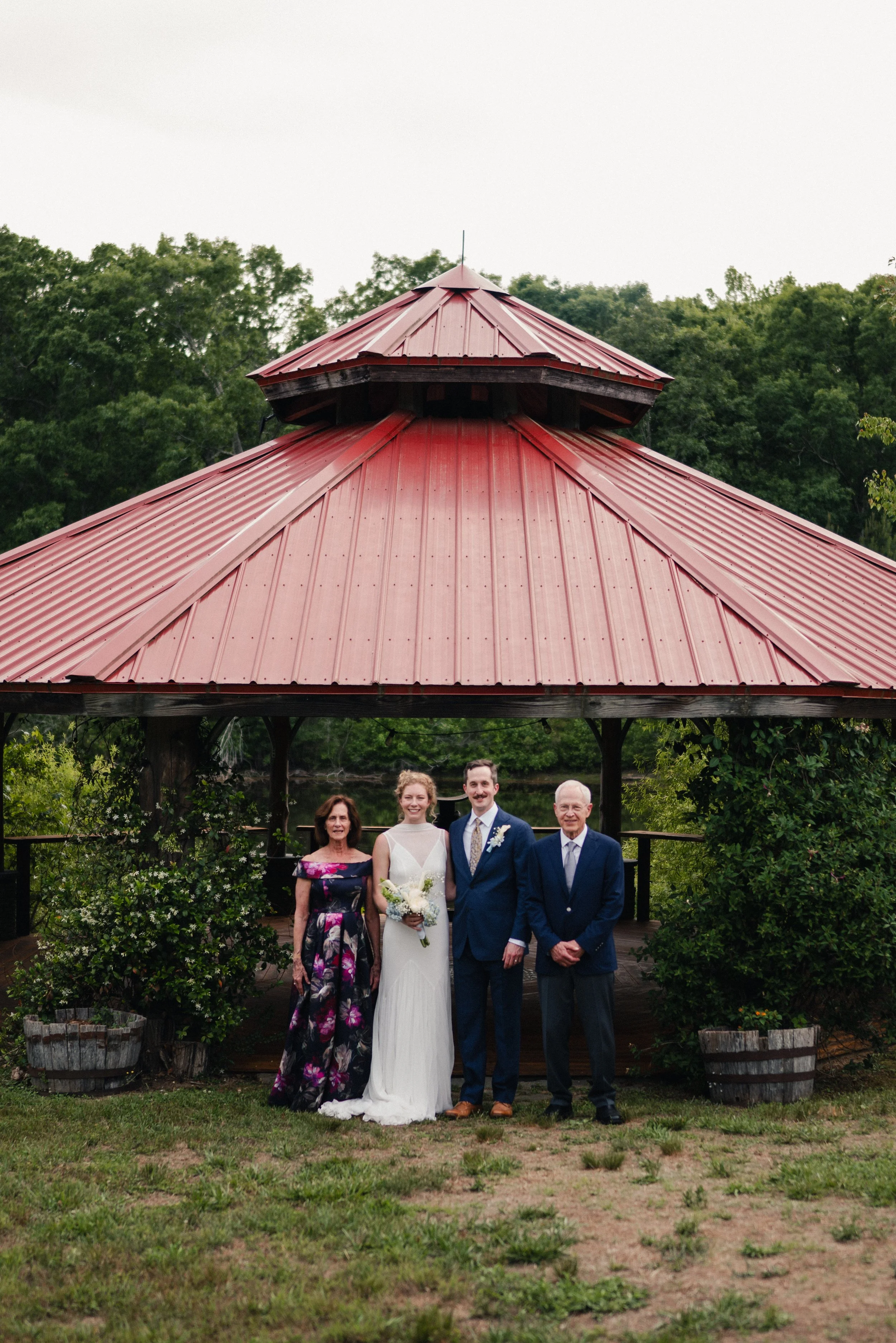 Group of four people standing outdoors under a red metal-roofed gazebo, with a woman in a wedding dress holding a bouquet, a man in a suit, and two older women, surrounded by greenery and potted plants.