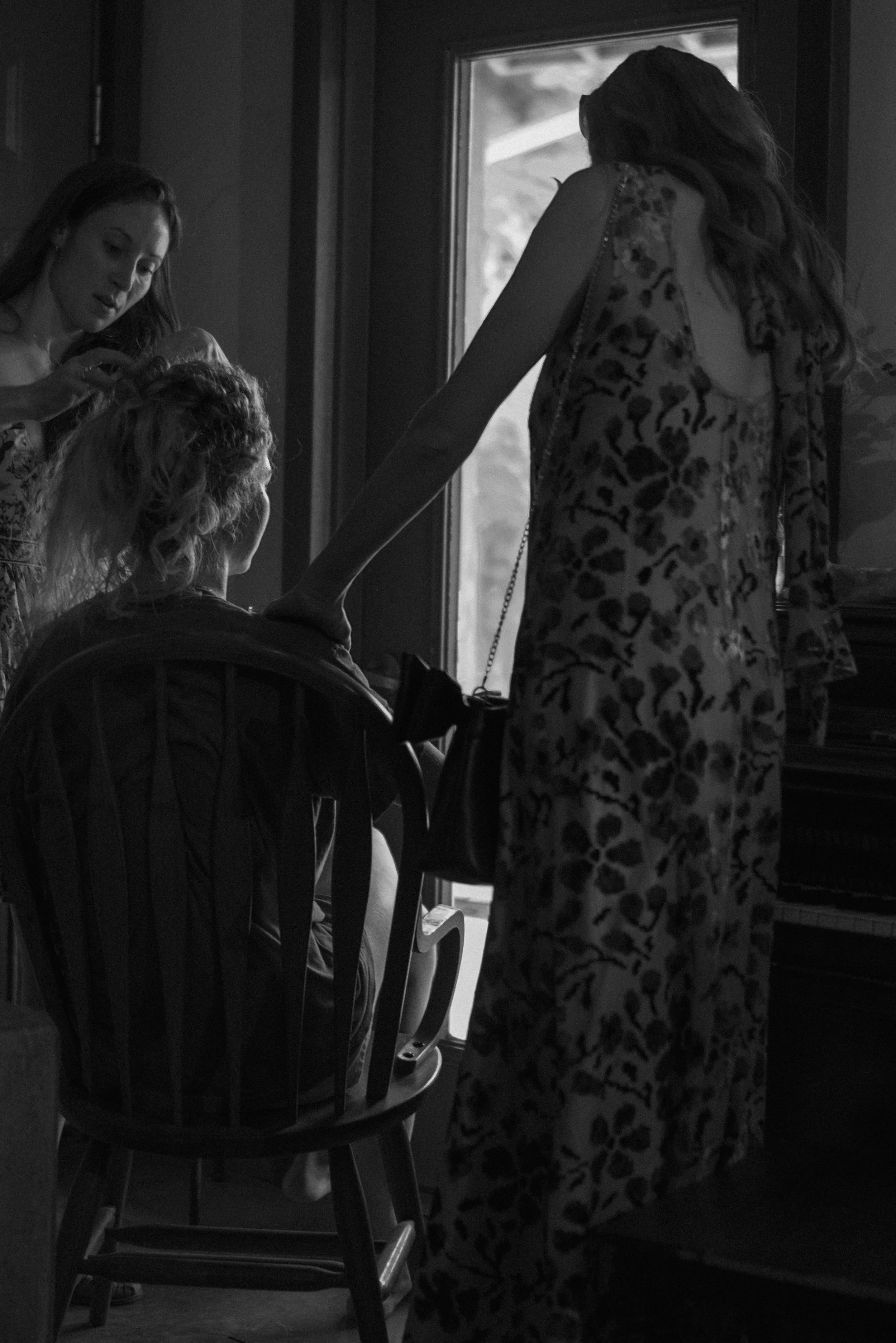 Three women are gathered in a room near a window, with one seated on a wooden chair and the other two standing. Two women are attending to the third woman, who is sitting and has curly hair. The standing woman on the right is wearing a patterned dres