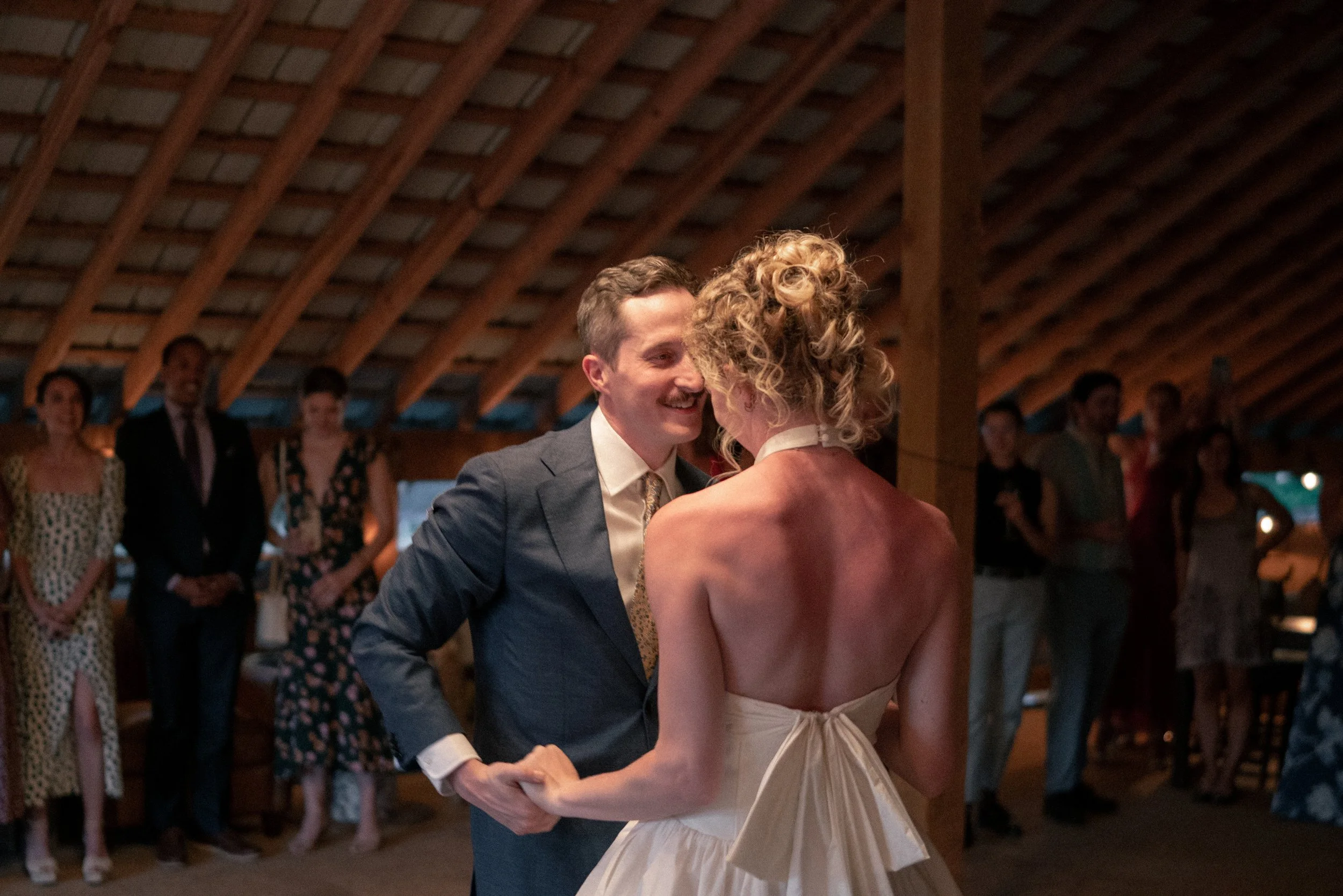 A man and a woman are dancing closely at a wedding reception, with guests watching in the background inside a wooden pavilion.
