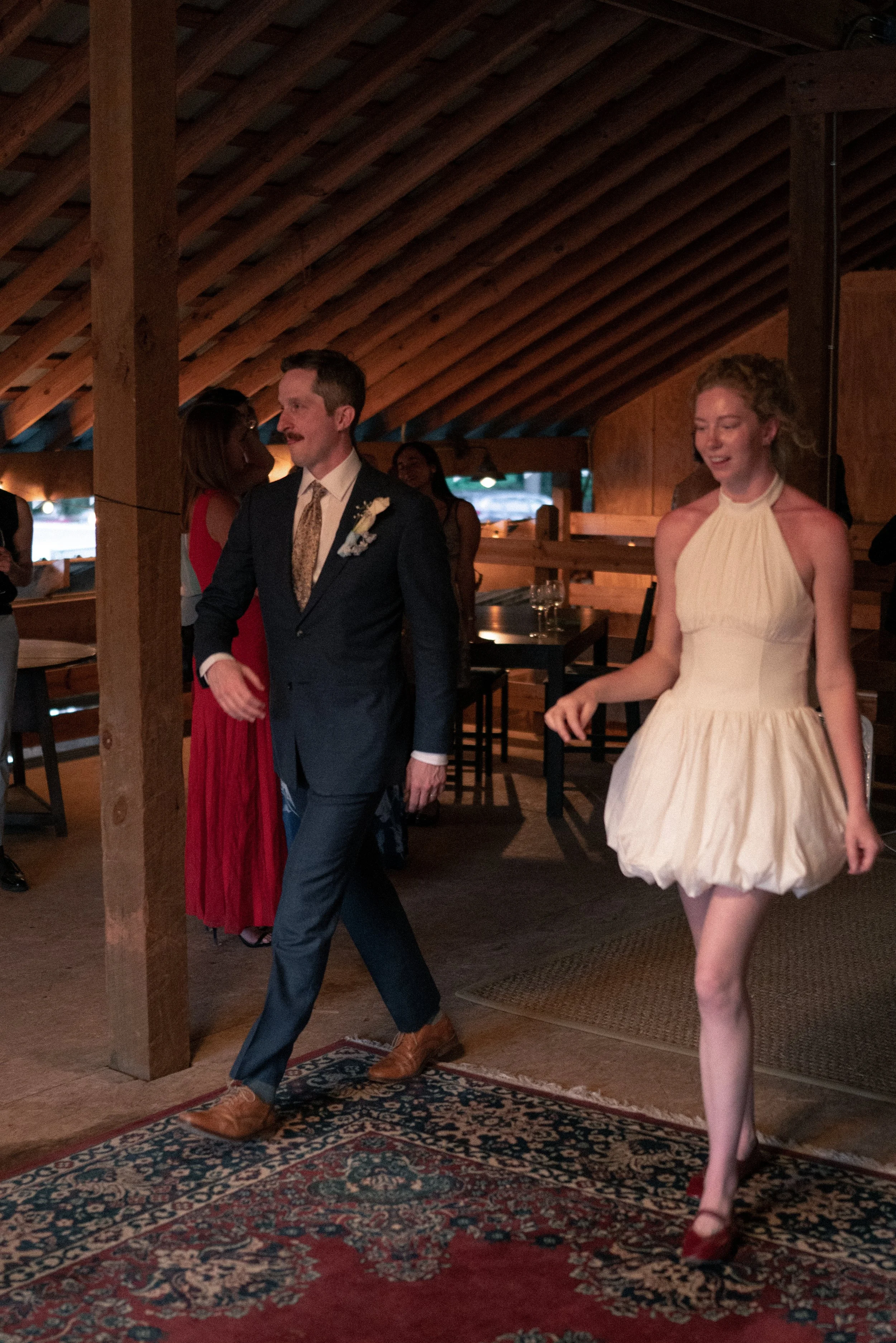 A man in a suit and a woman in a cream dress dancing at a celebration in a wooden venue. Other guests are visible in the background, and the setting appears warm and festive.