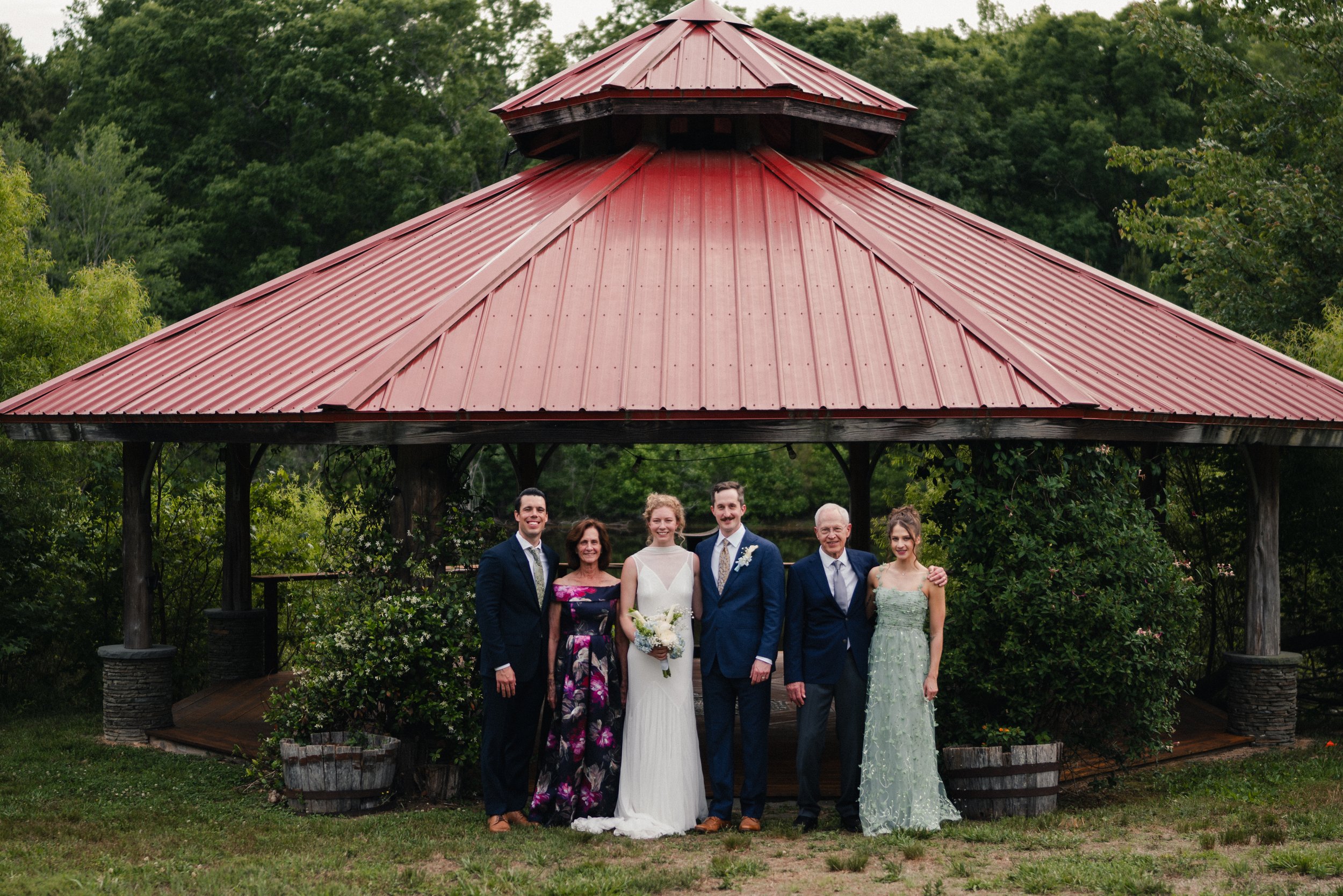 Group of people dressed in formal attire, standing outdoors in front of a pavilion with a red-metal roof, lush green trees in the background, celebrating a wedding.