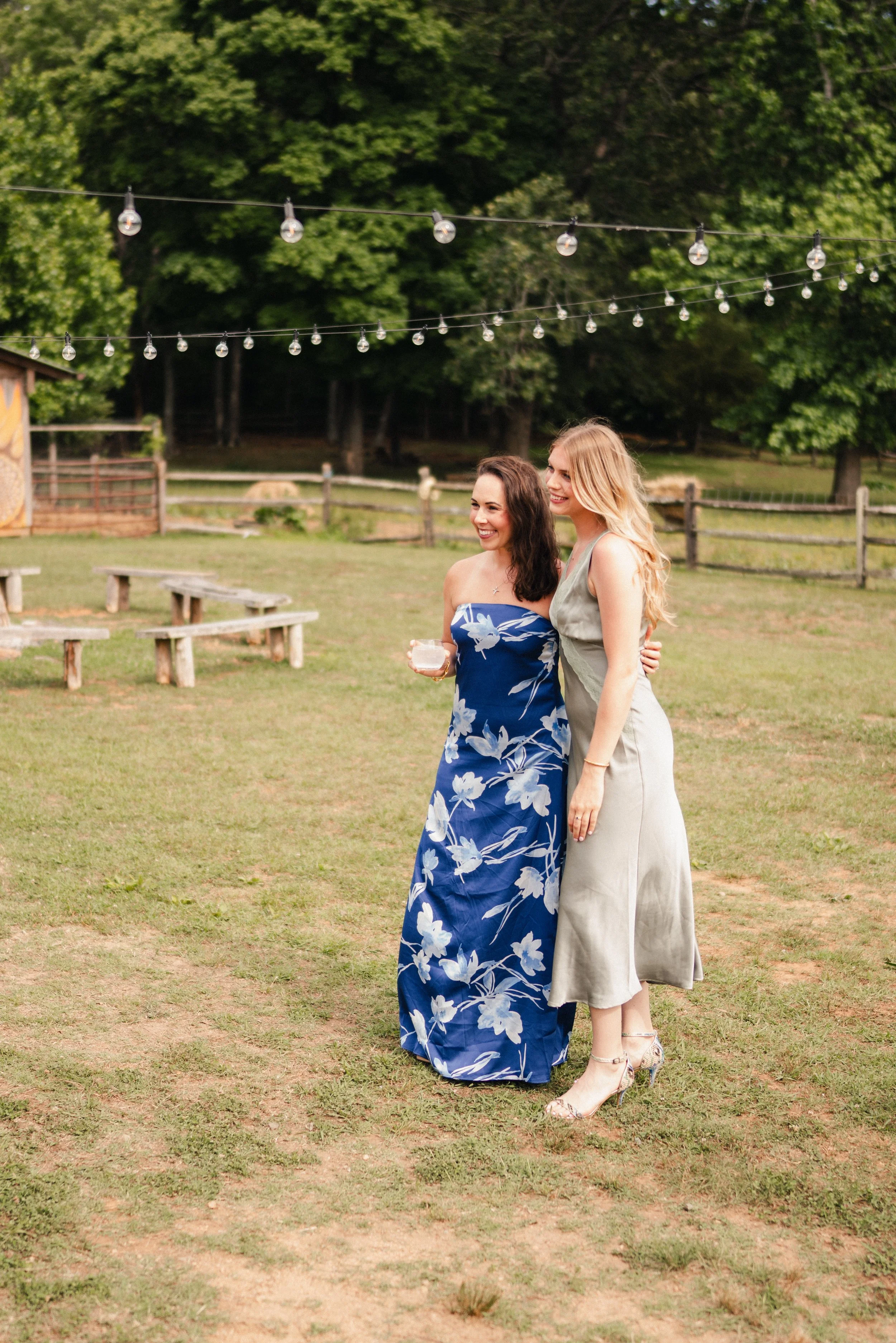 Two women standing close together outdoors, smiling, under string lights, with trees and a wooden fence in the background.