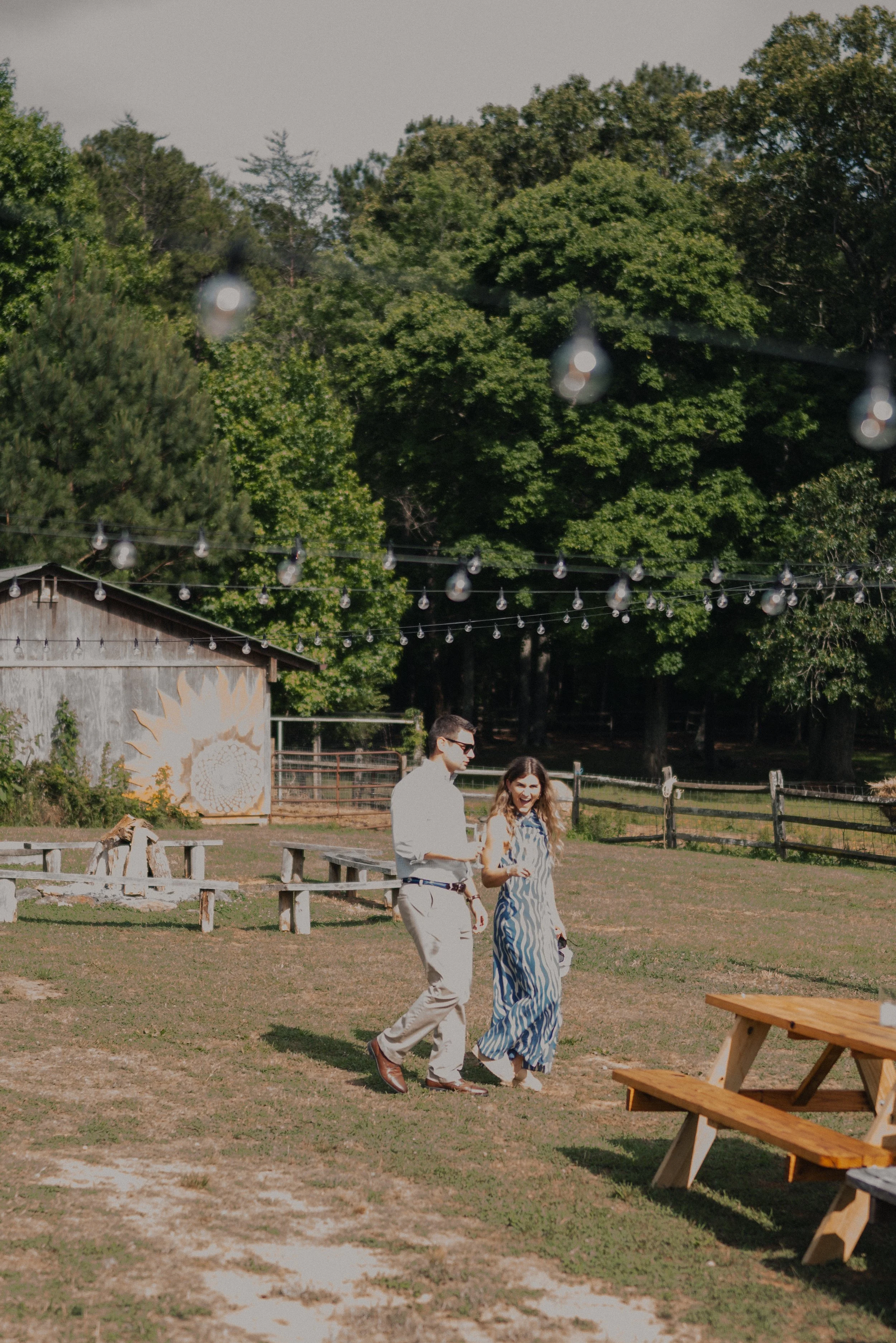 Two people walking together in a grassy outdoor area with trees, string lights hanging overhead, a wooden building, and picnic tables.